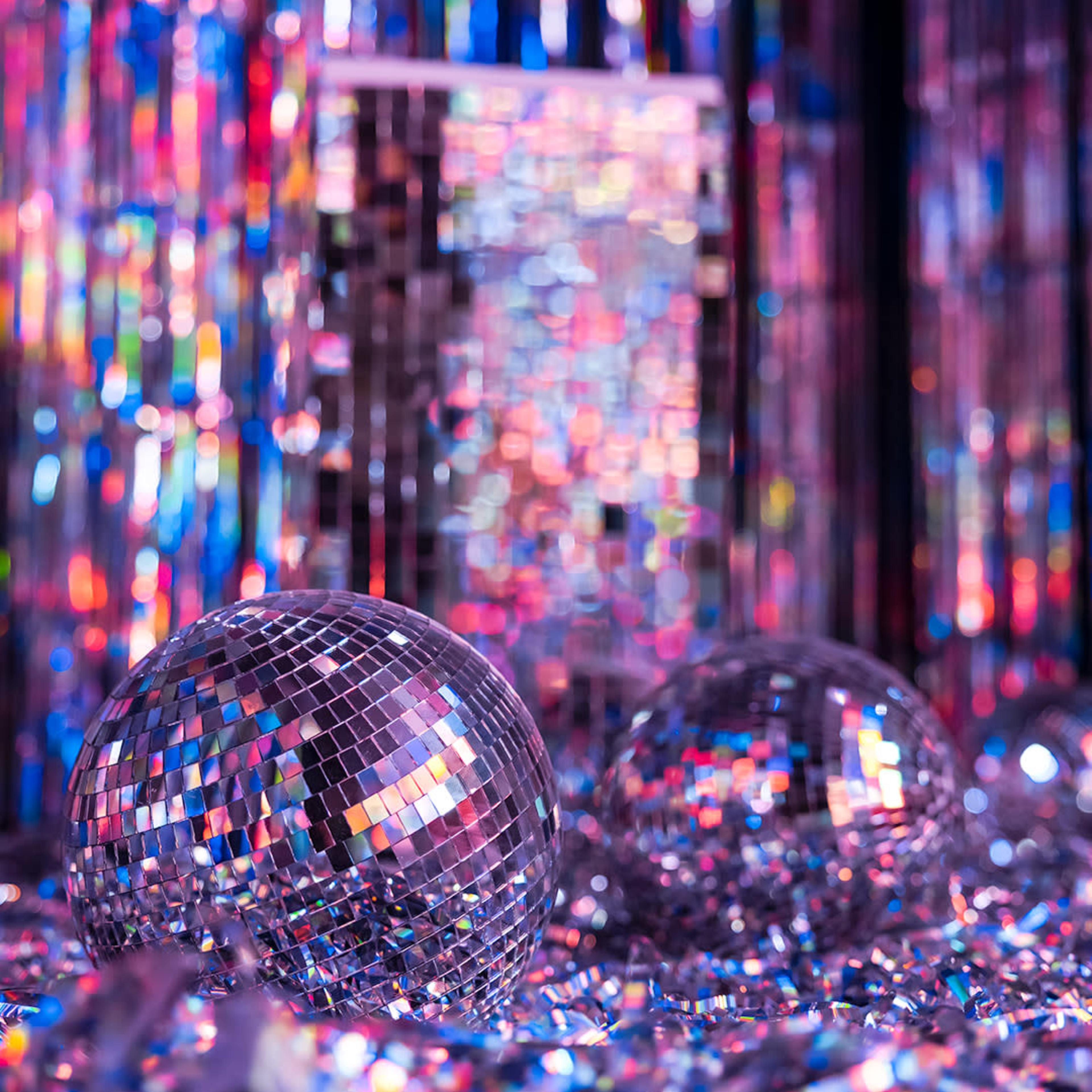 Two reflective disco balls surrounded by scattered pieces of colorful shimmering material against a backdrop of sparkling curtains.