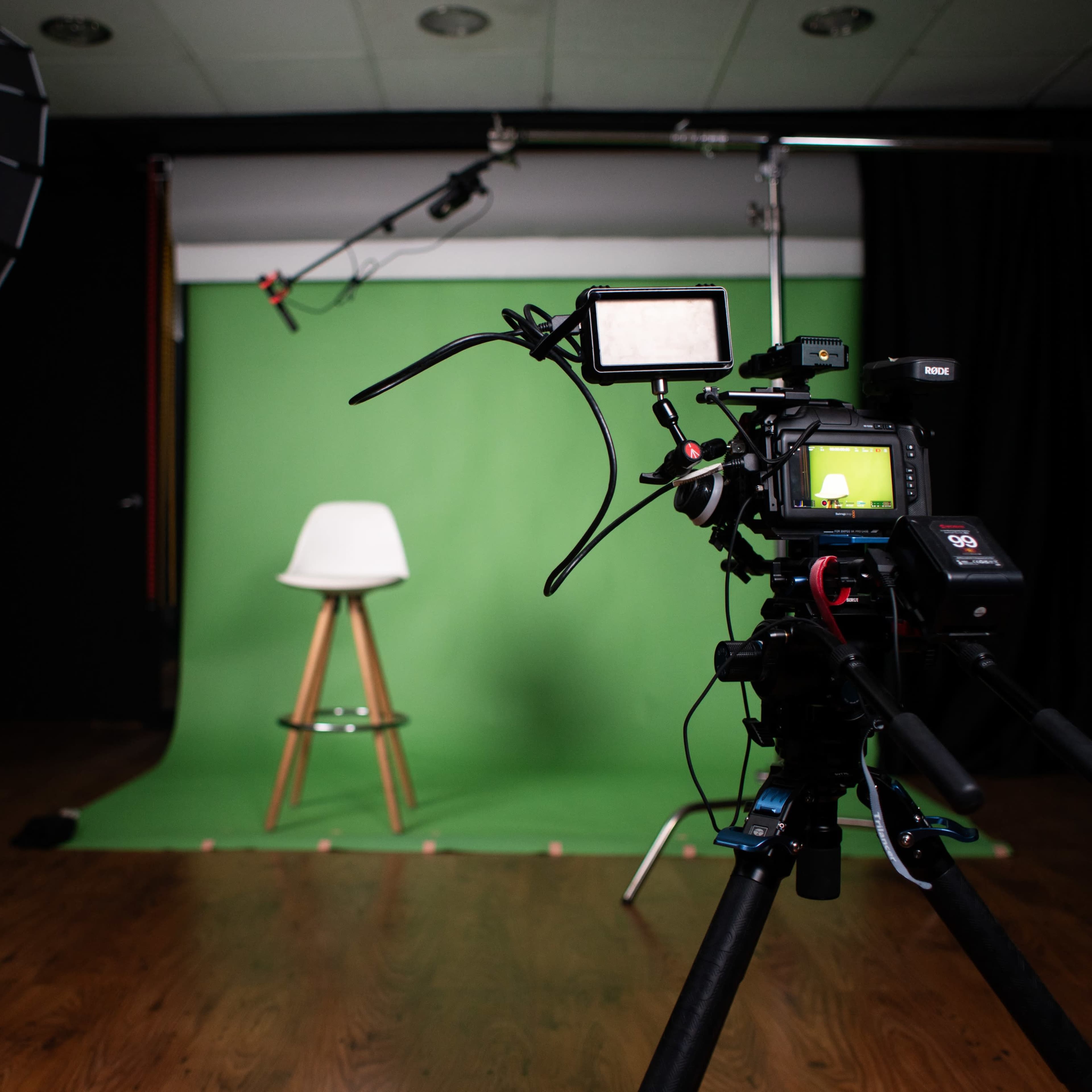 A camera is positioned in front of a green screen with a white stool placed on a wooden floor in a studio setting.