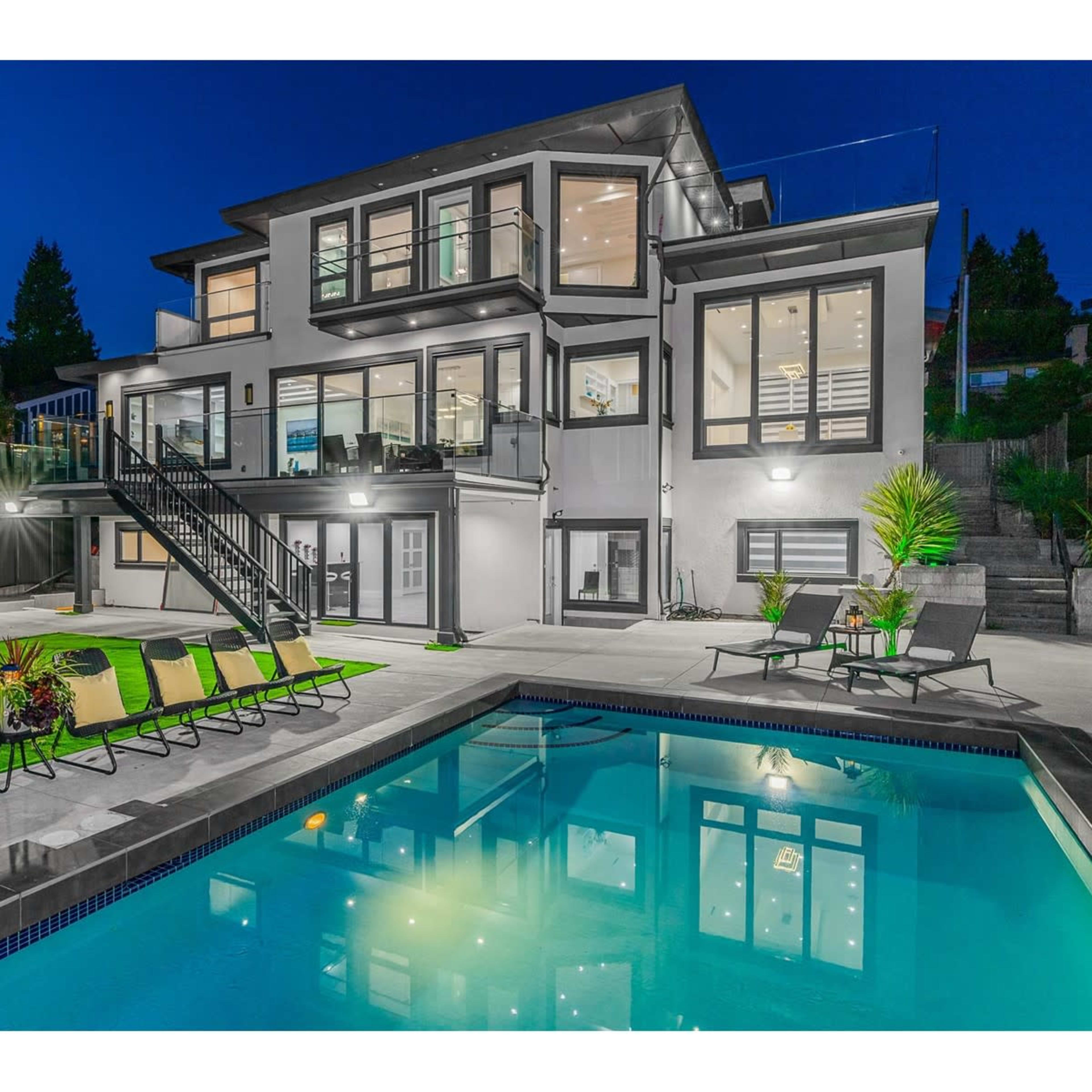 A modern two-story house with large windows overlooks a rectangular pool surrounded by patio chairs and a grassy yard at night.