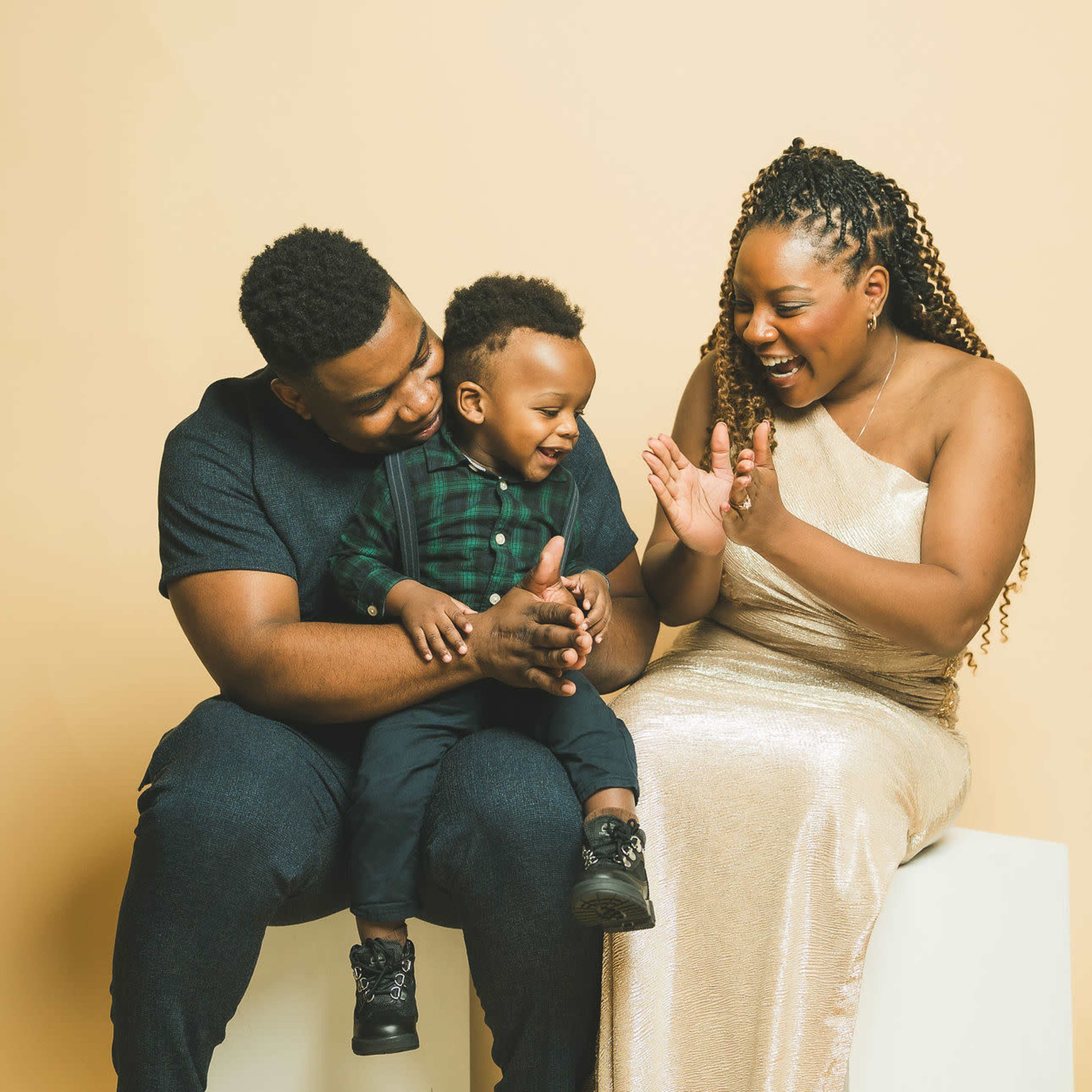 A family of three, seated on a white cube against a beige background, shares a joyful moment as they play and clap together.