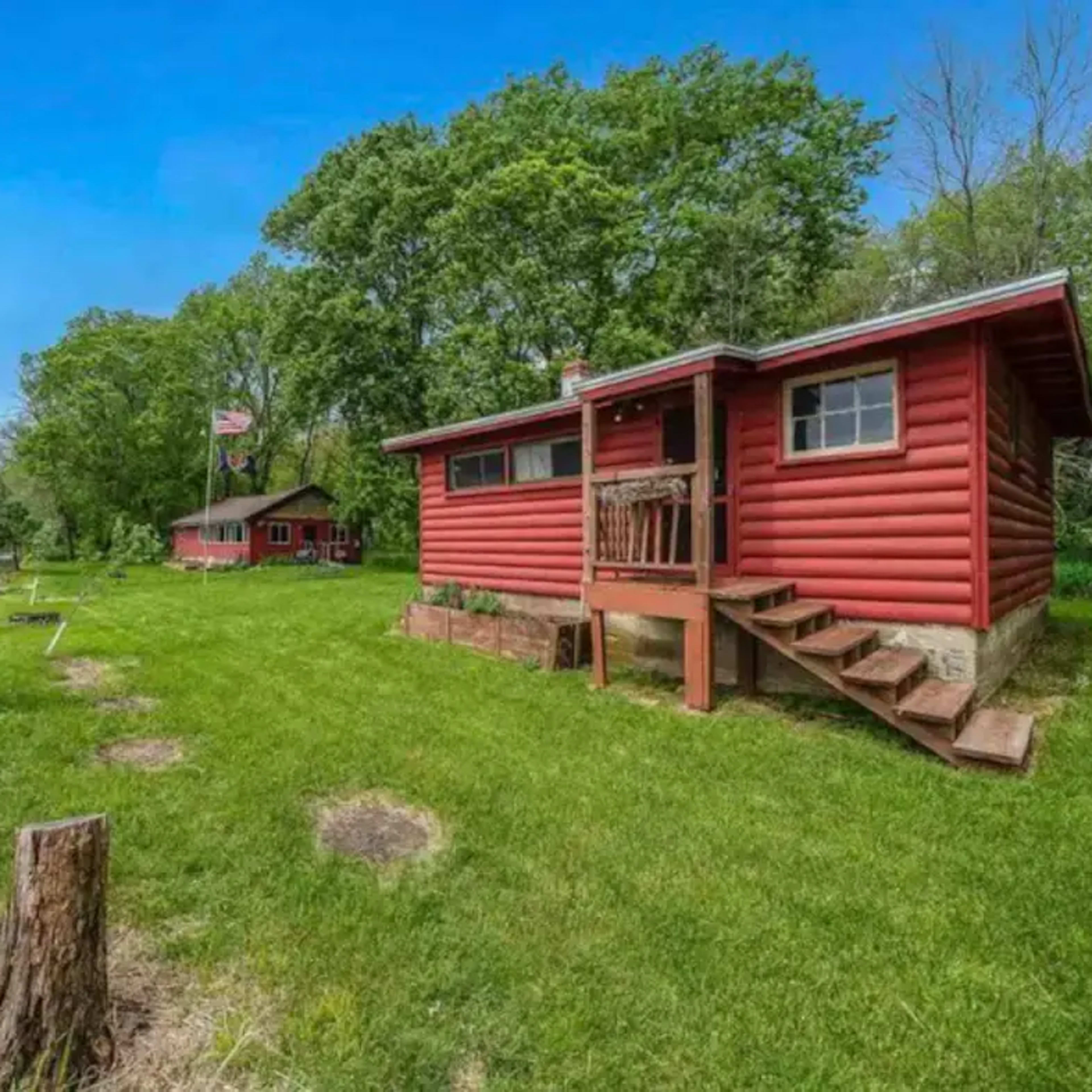A red log cabin with a porch is situated near a lake, surrounded by trees and a grassy area.