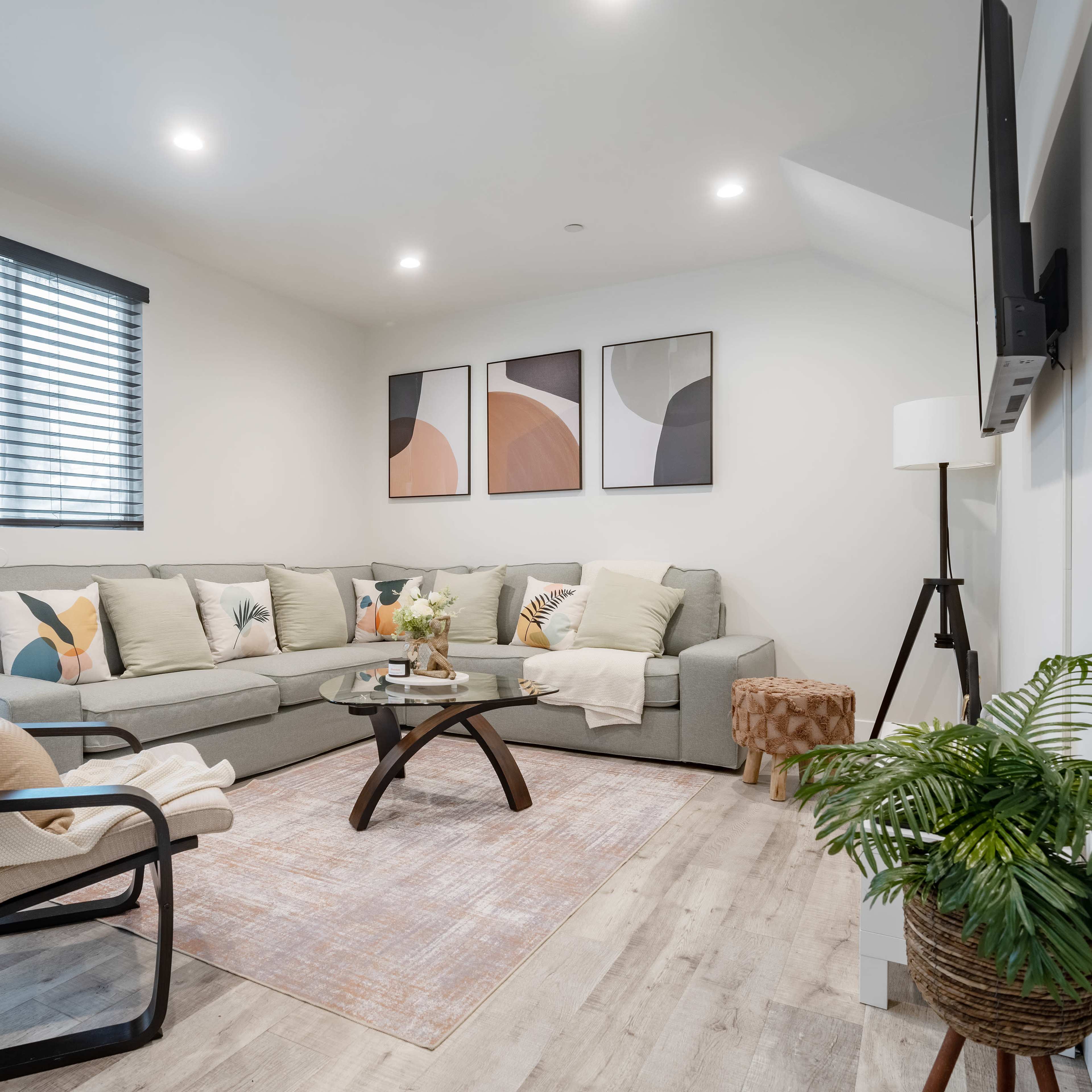 A modern living room features a gray sectional sofa, a wooden coffee table, and tropical-themed decor, with natural light streaming through the window.