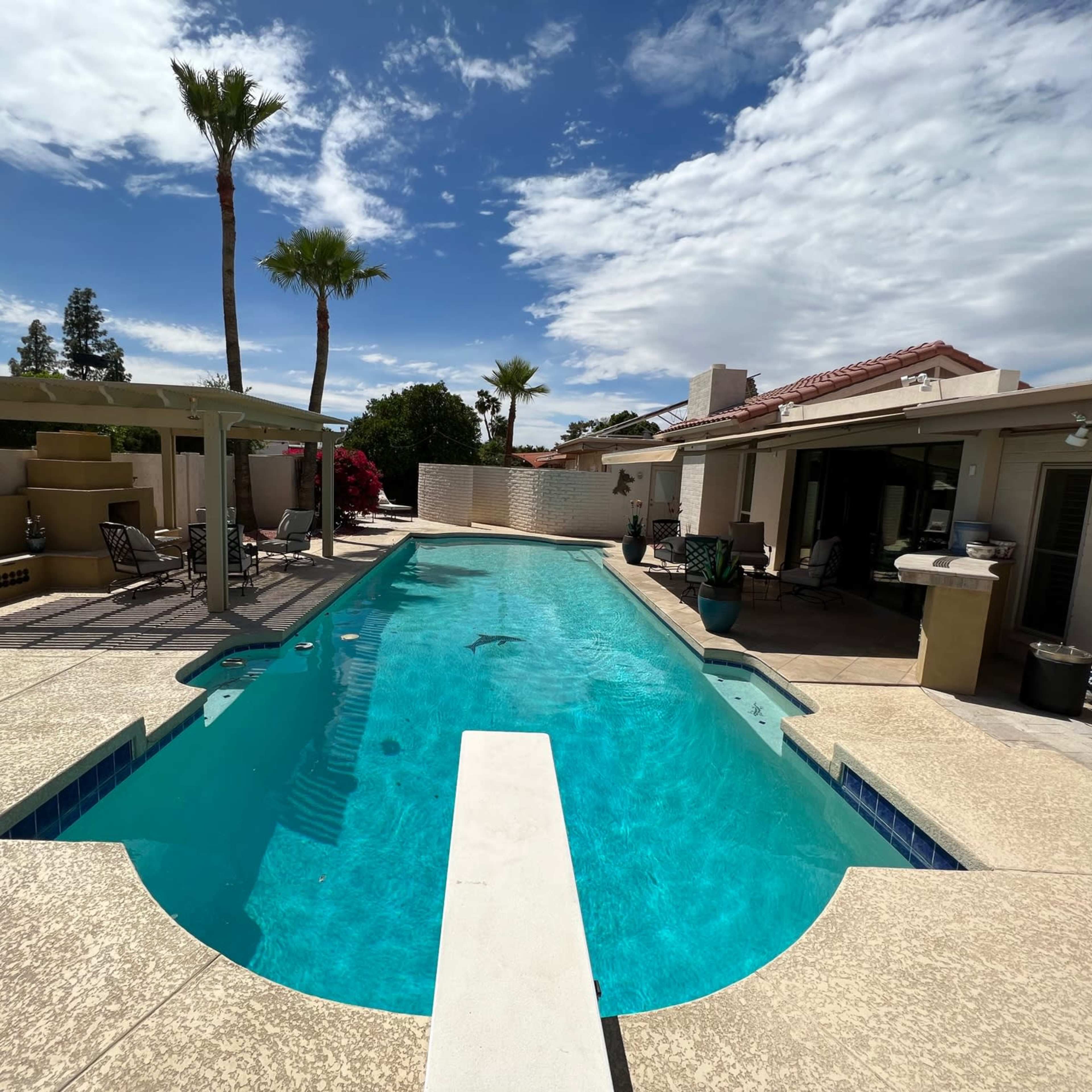 A swimming pool surrounded by palm trees and a patio area with outdoor furniture and a barbecue grill.