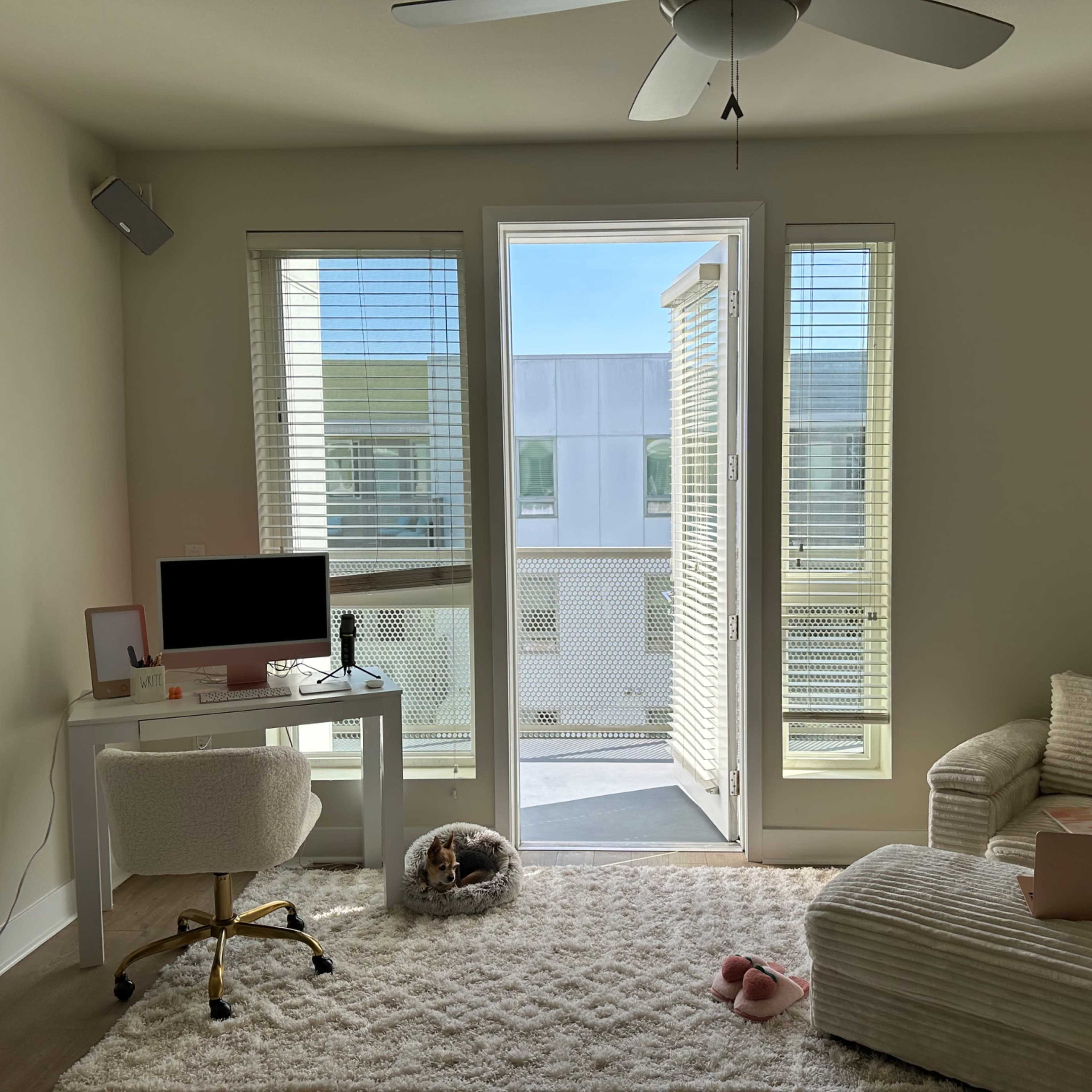 A light-colored room with a desk featuring a computer, a chair, and a cozy sofa, along with large windows leading to a balcony.