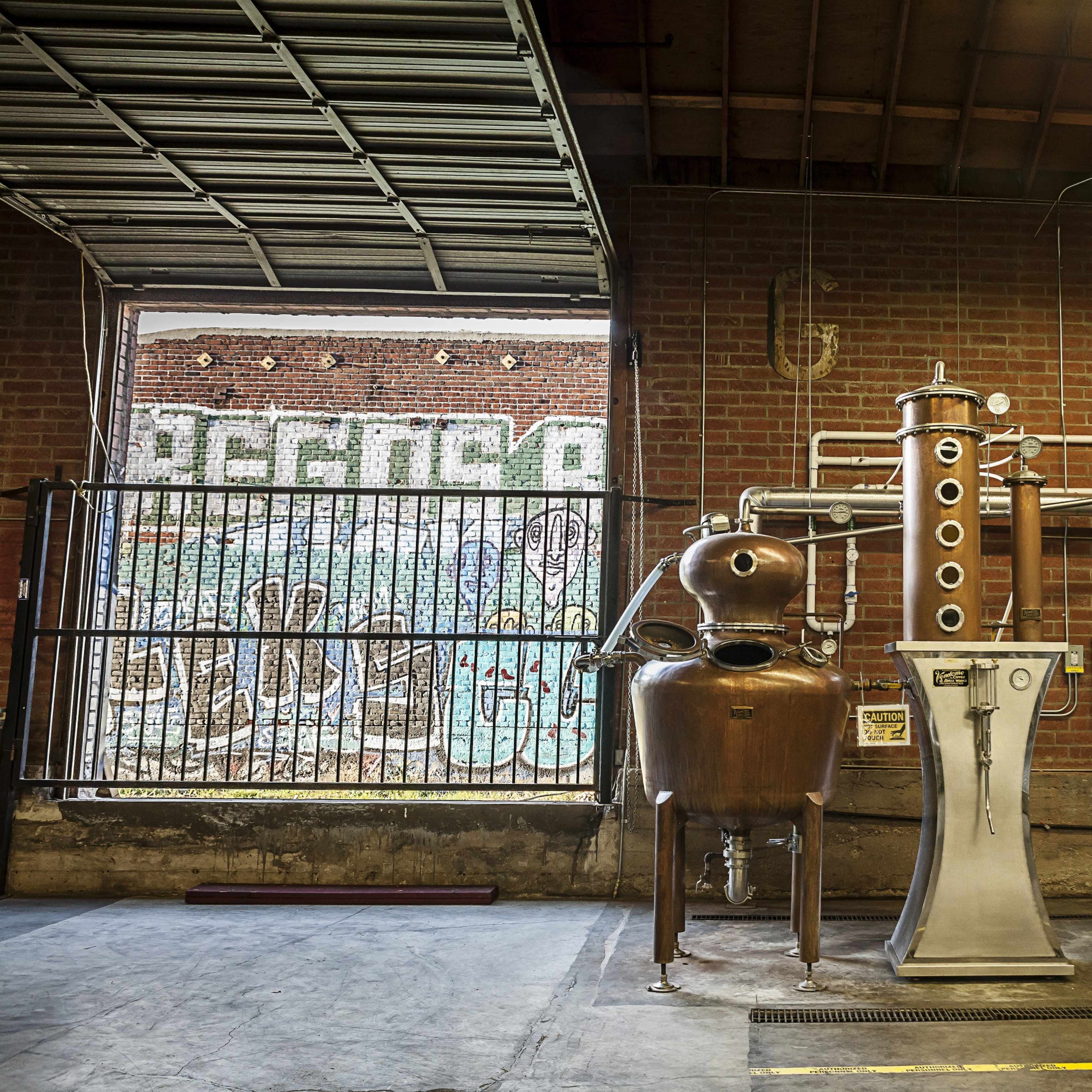 A distillation apparatus made of copper and stainless steel set against a brick wall with graffiti.