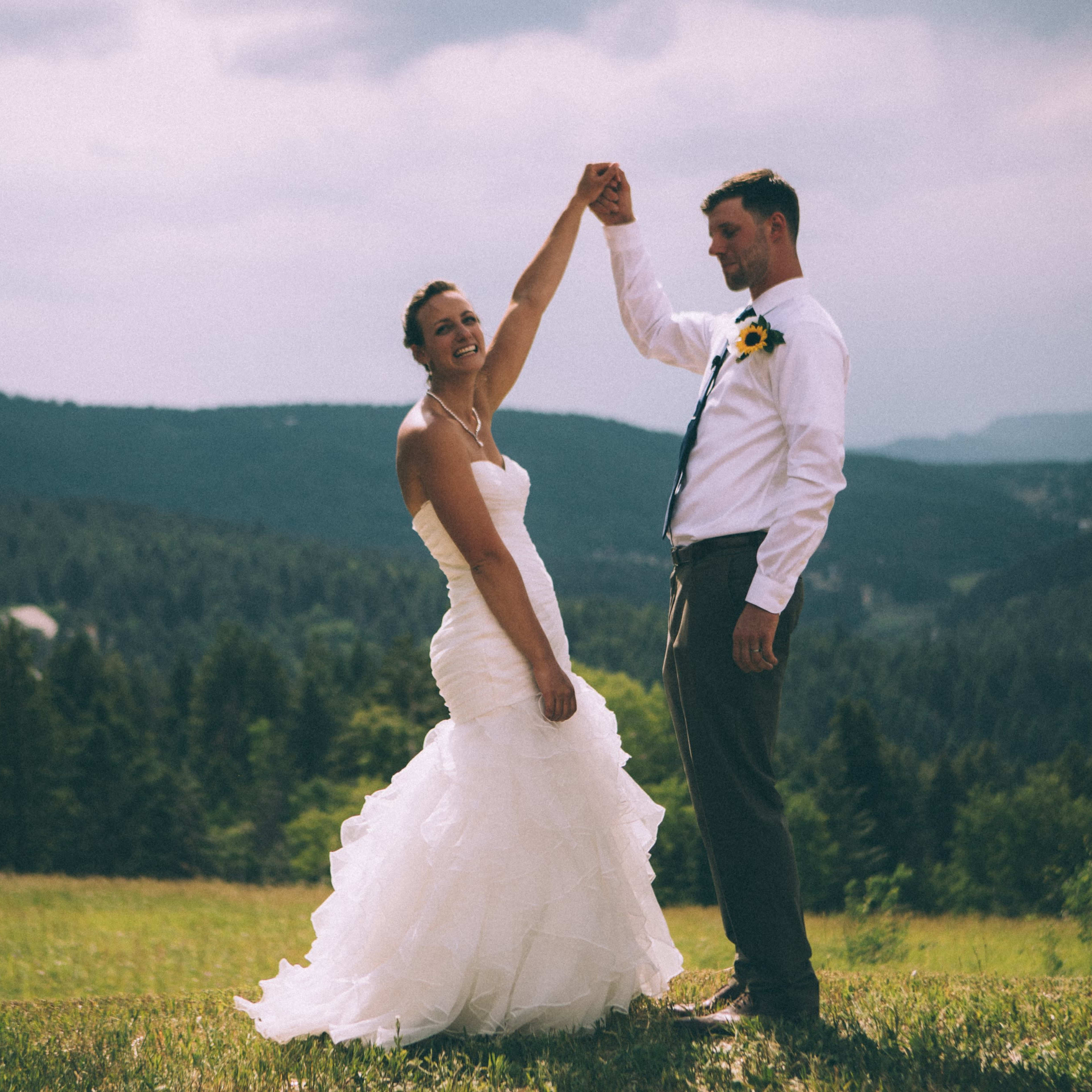 A bride and groom dance outdoors, holding hands while surrounded by a forested landscape and distant mountains.