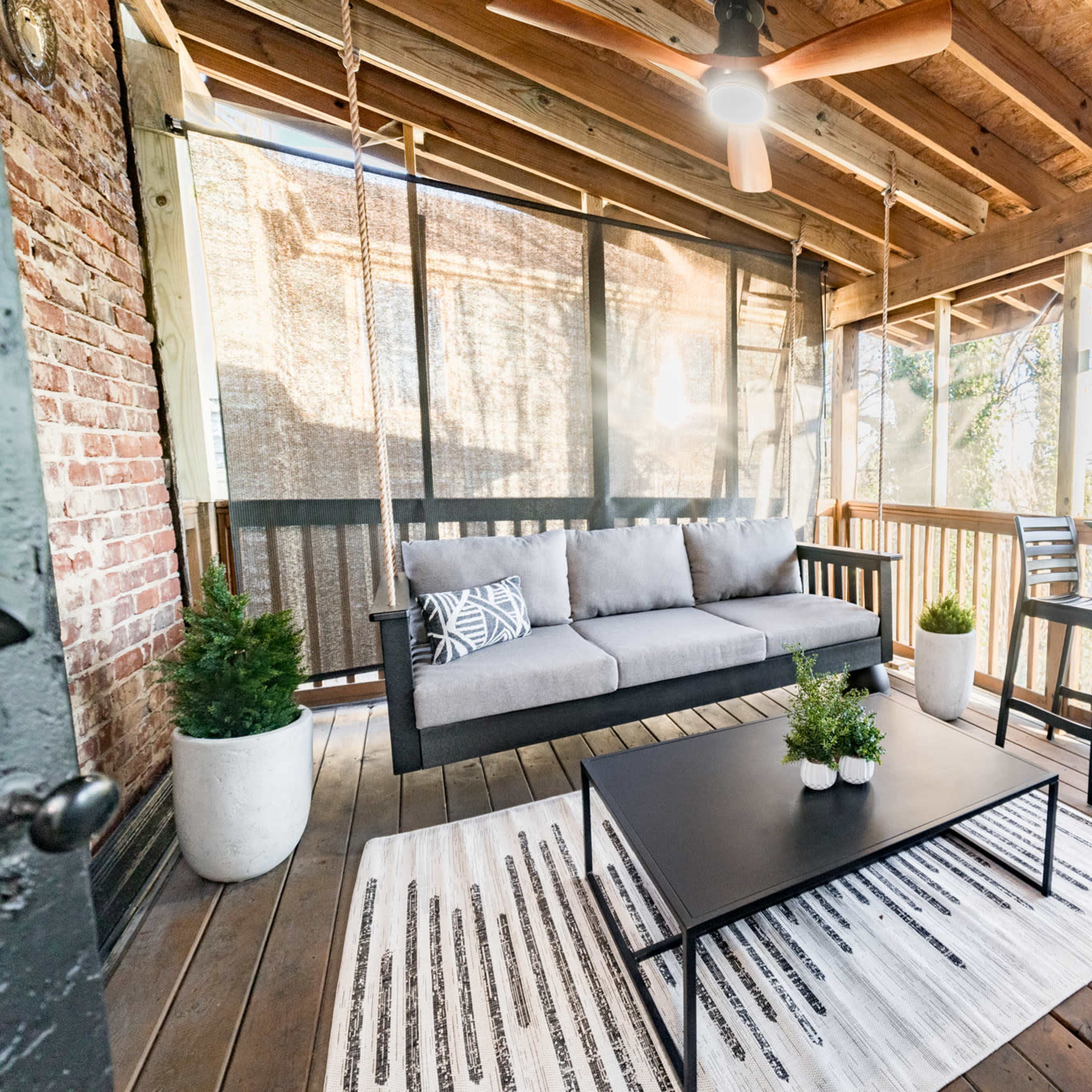 A covered outdoor patio features a gray sofa, a black coffee table, and two high chairs, surrounded by greenery and wooden beams.