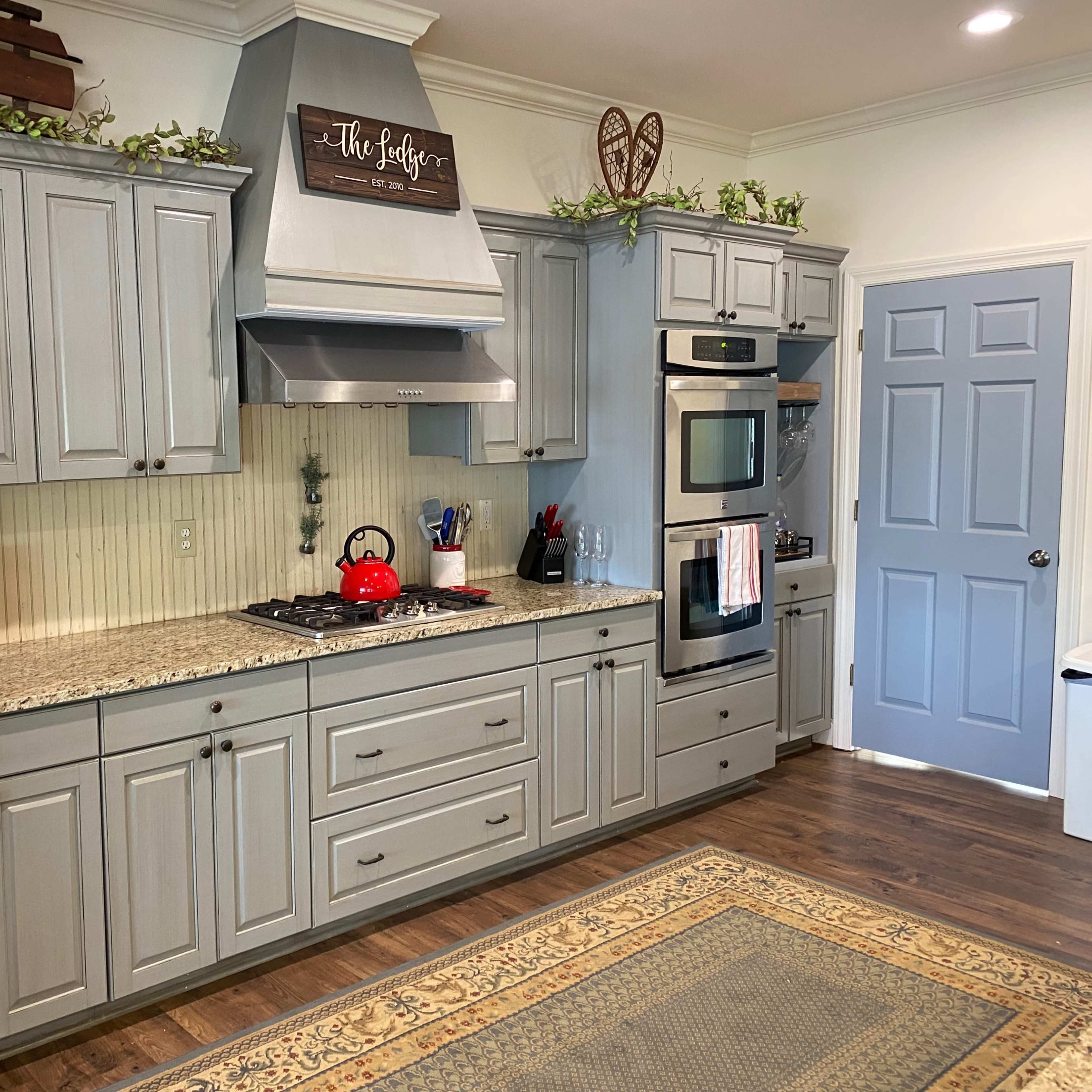 A modern kitchen with gray cabinets, a stainless steel refrigerator, a gas stove with a red kettle, and a decorative rug on the wooden floor.