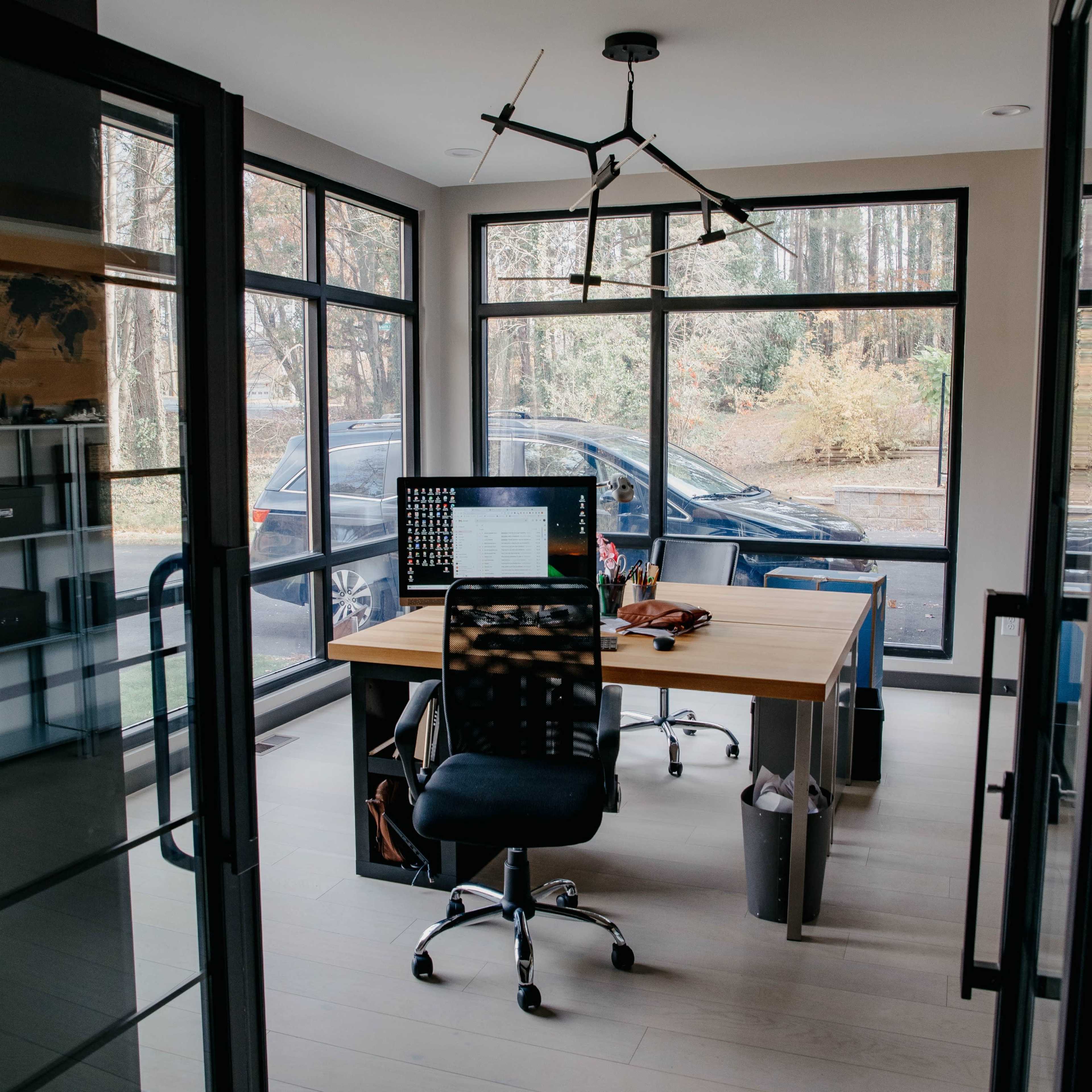 A modern home office with a large window, a wooden desk with a computer, and a view of a parked car outside.