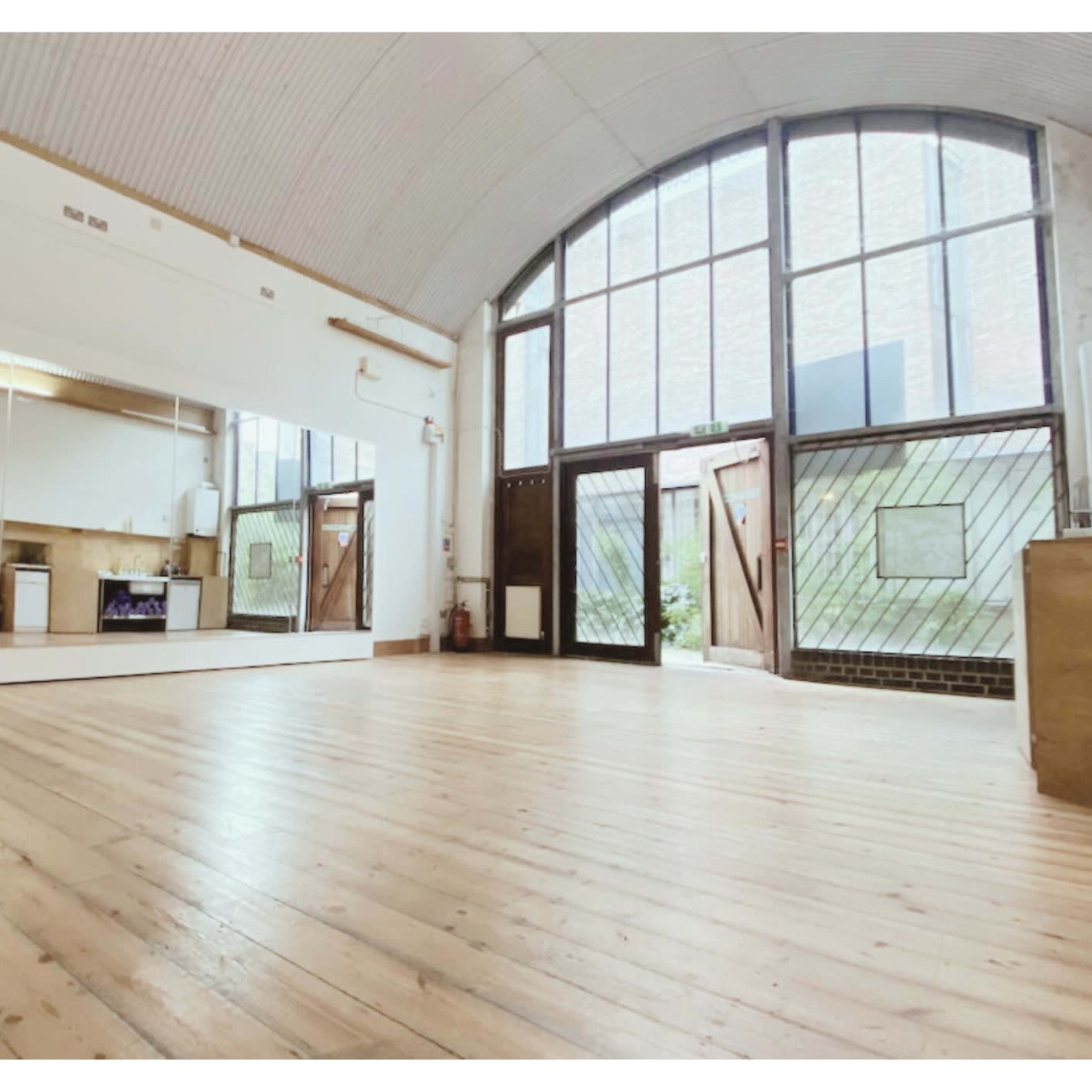 A spacious room with wooden flooring, large windows, and mirrors on one wall, leading to an outdoor area.