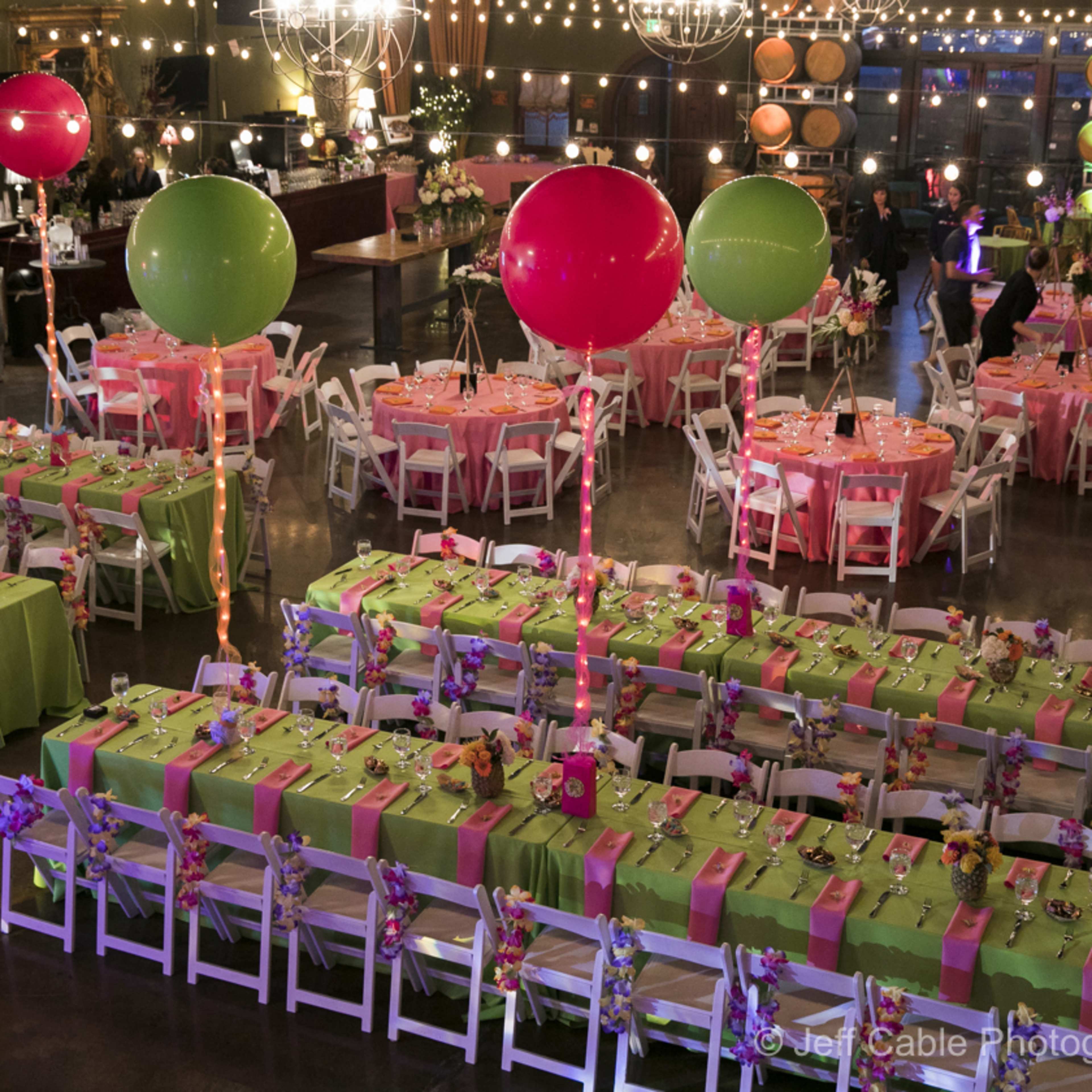 A large banquet hall set up for an event, featuring rows of tables adorned with colorful tablecloths and decorations, balloons, and string lights overhead.