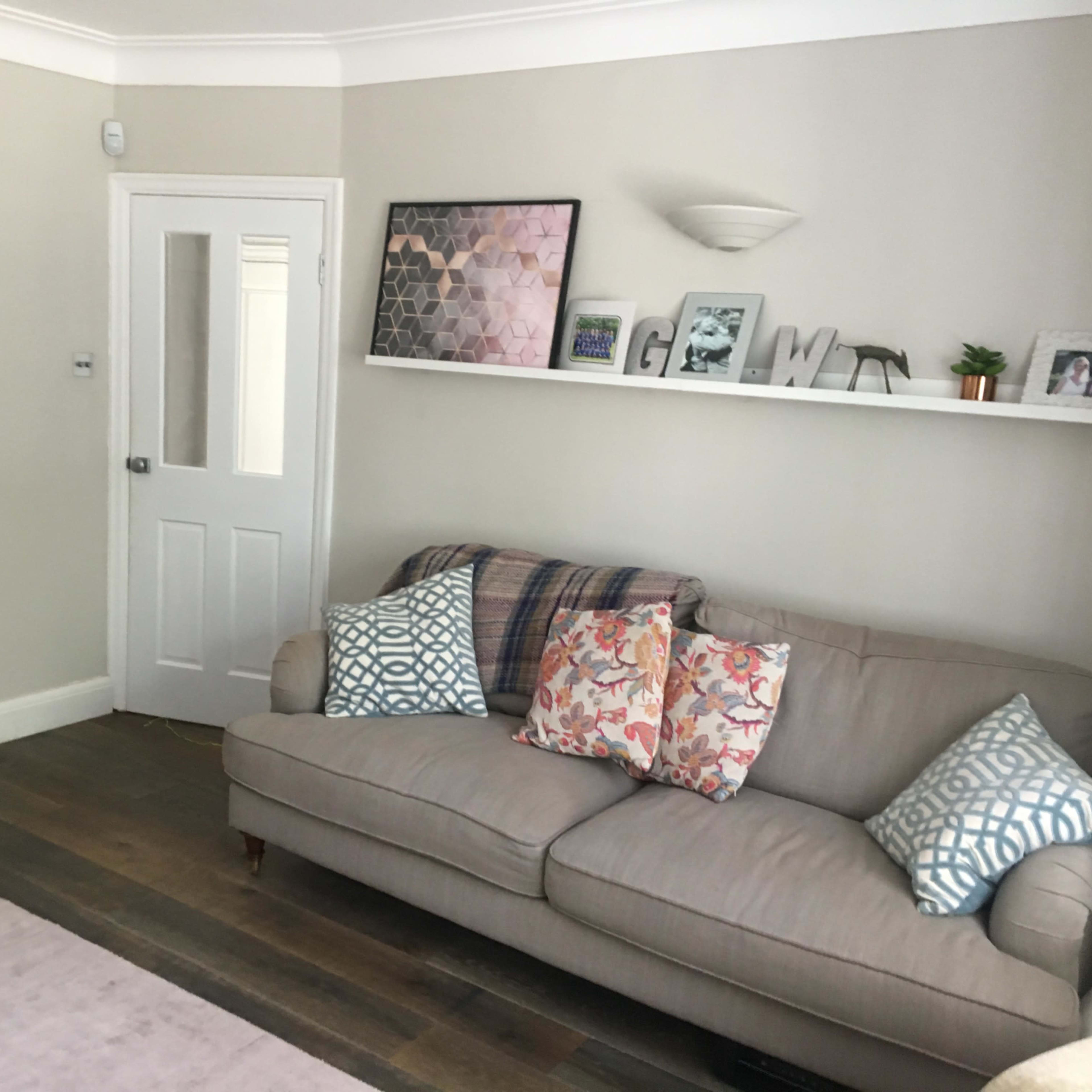 A light-colored living room features a beige sofa with patterned cushions, a wooden TV stand, and a wall shelf displaying decorative items and photos.