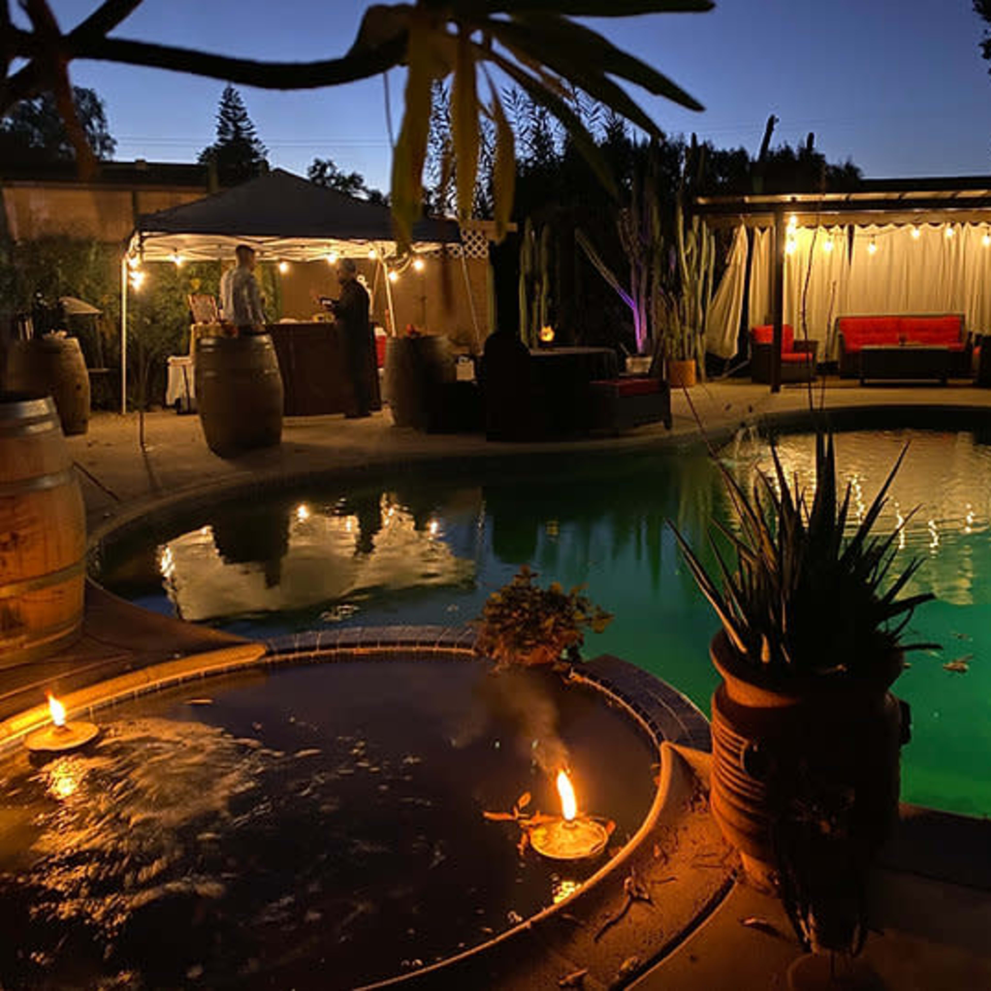 A pool area at night features a lit water feature beside the pool, with surrounding greenery and a lounge area illuminated by soft lighting.