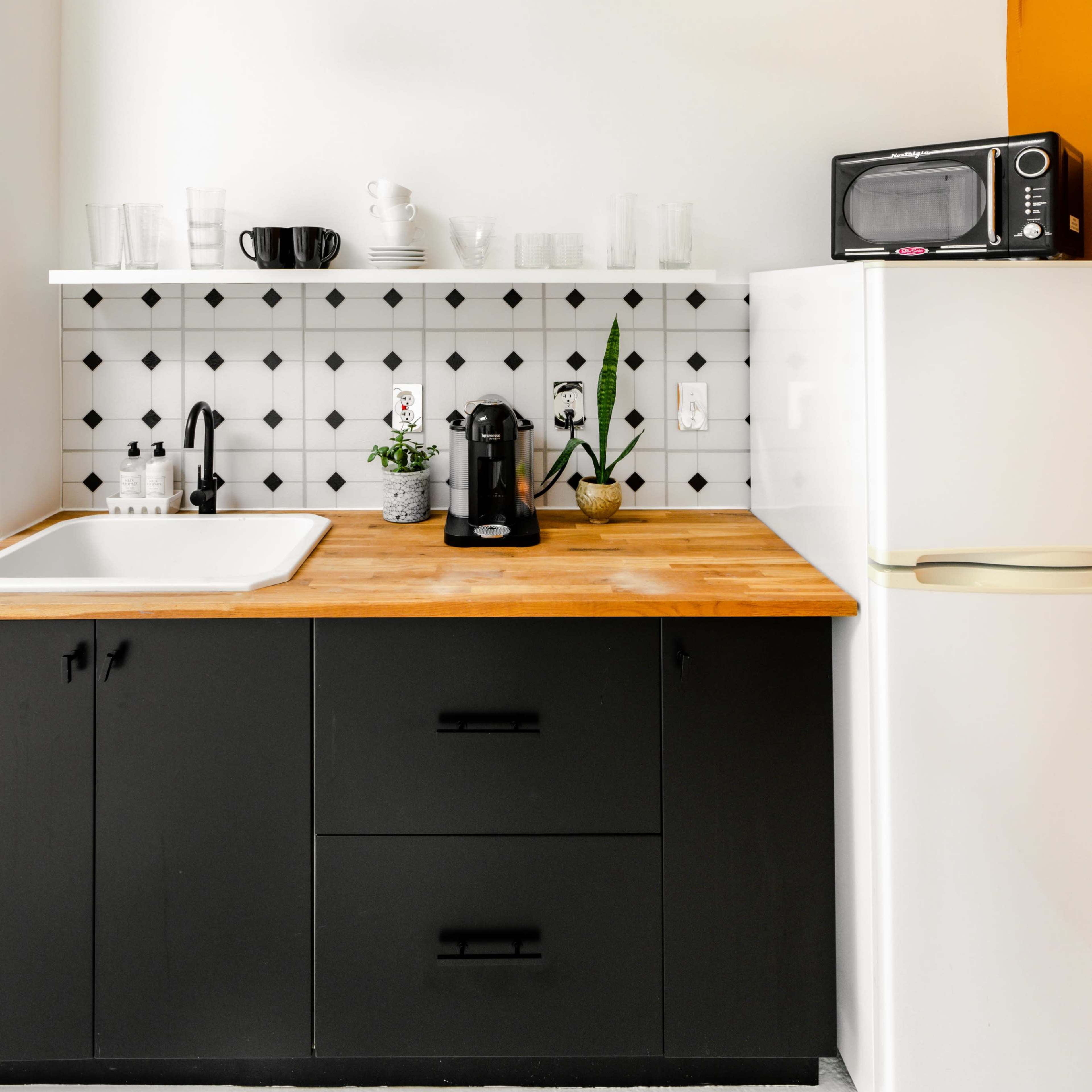 A modern kitchen with black cabinetry, a wooden countertop, a white tiled backsplash, and appliances including a microwave and coffee maker.