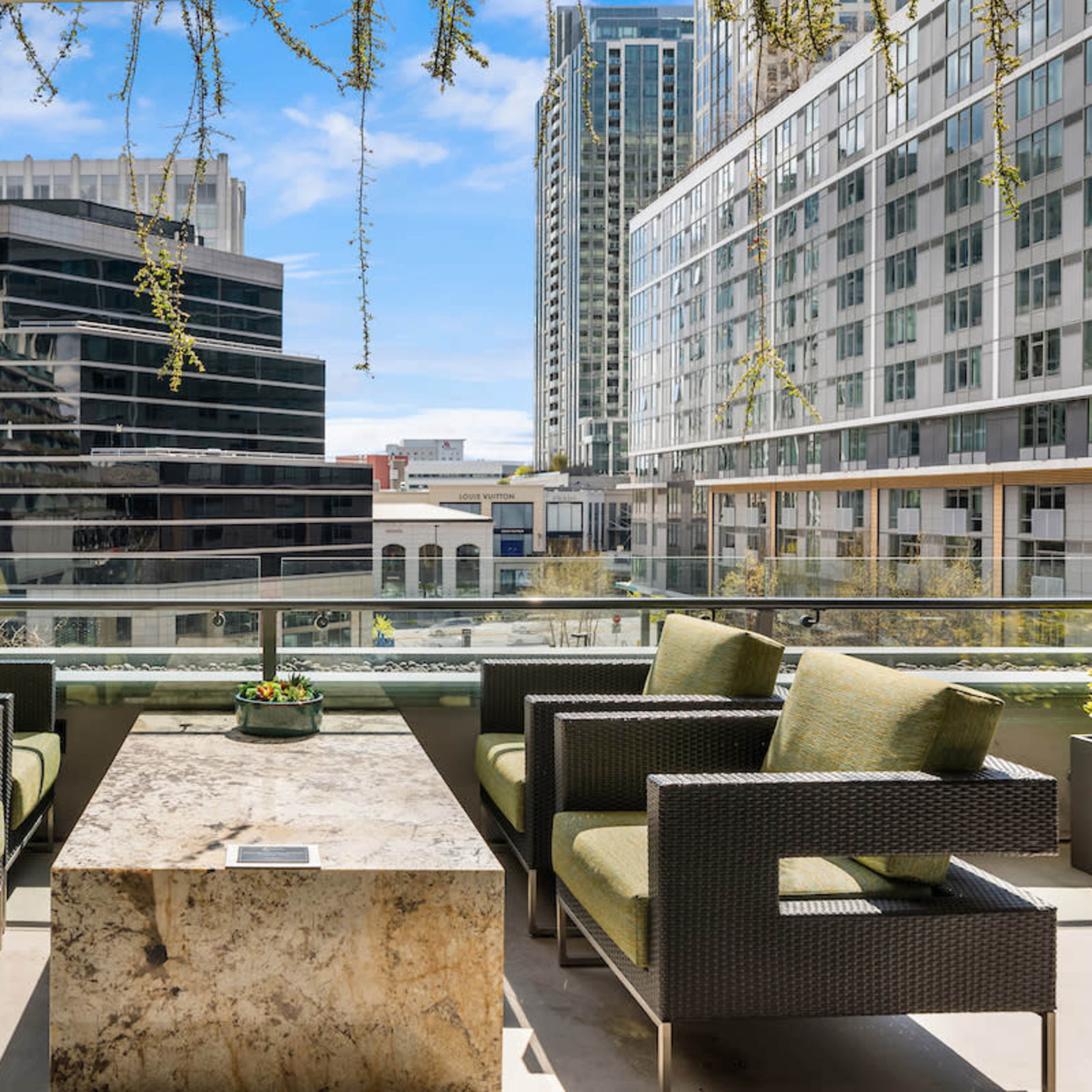 A balcony space features four green-upholstered chairs arranged around a stone table, overlooking modern buildings and greenery in an urban setting.