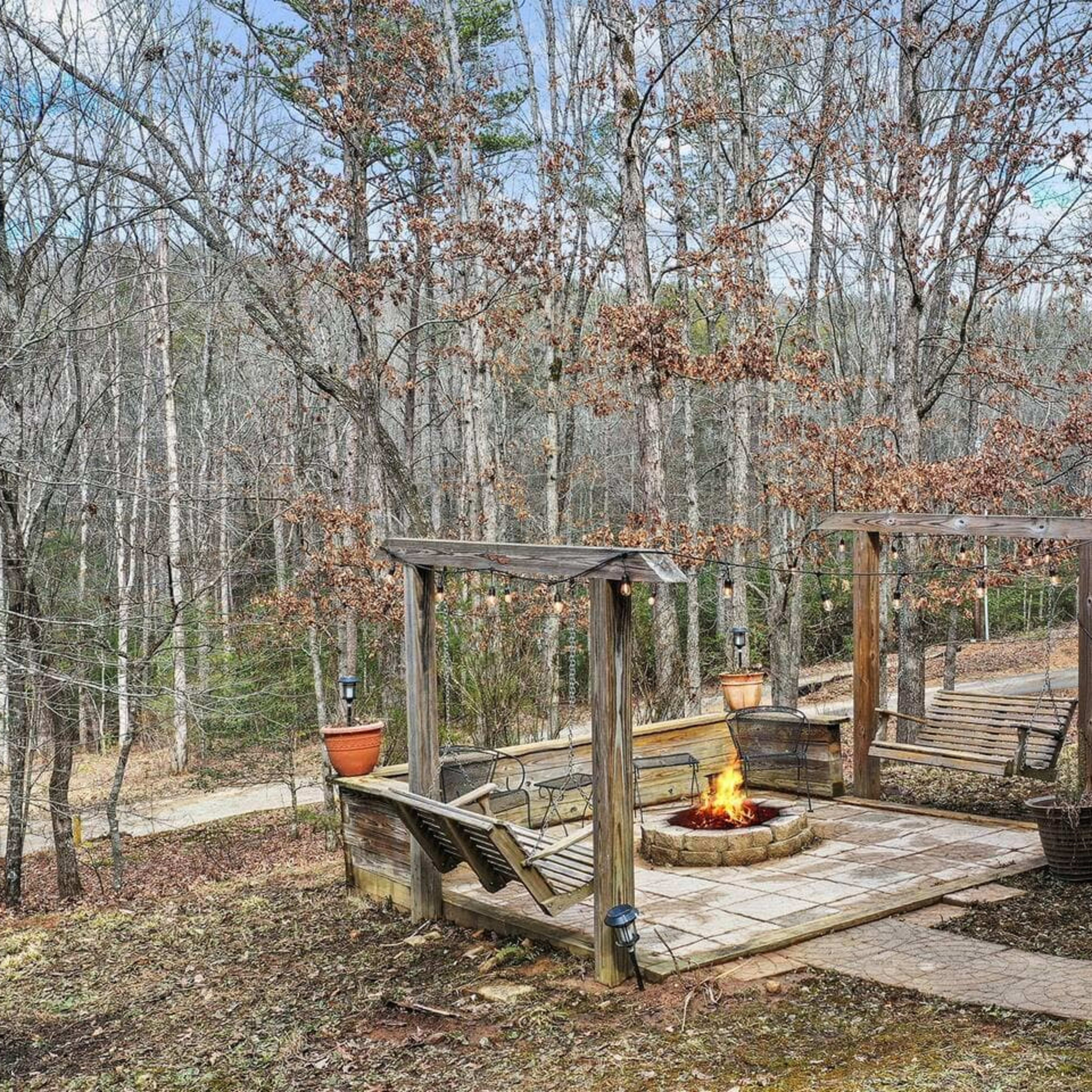 A rustic fire pit area with wooden benches surrounded by trees and a path in the background.