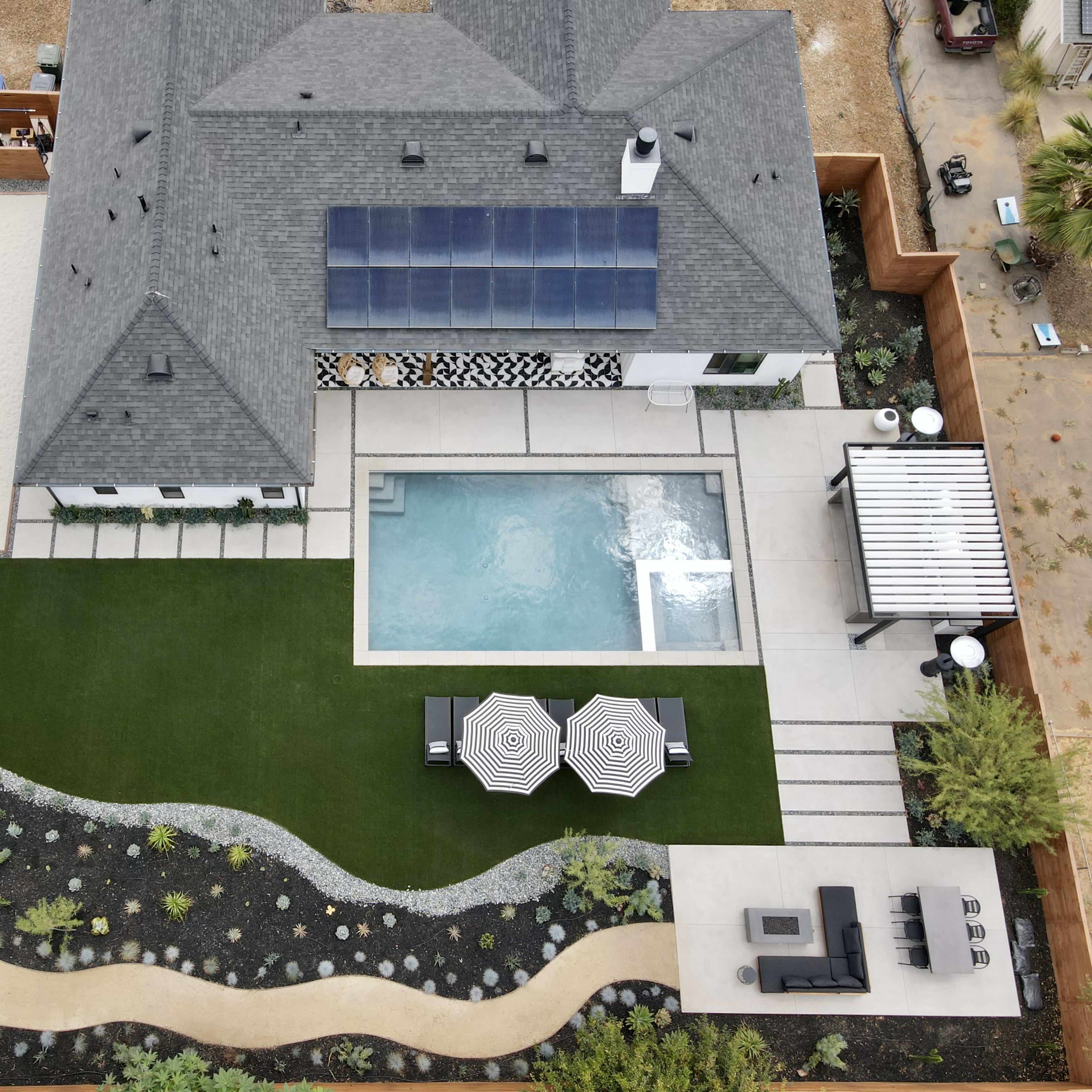 A landscaped backyard featuring a swimming pool, a patio area with two striped umbrellas, a putting green, and a house with solar panels on the roof.