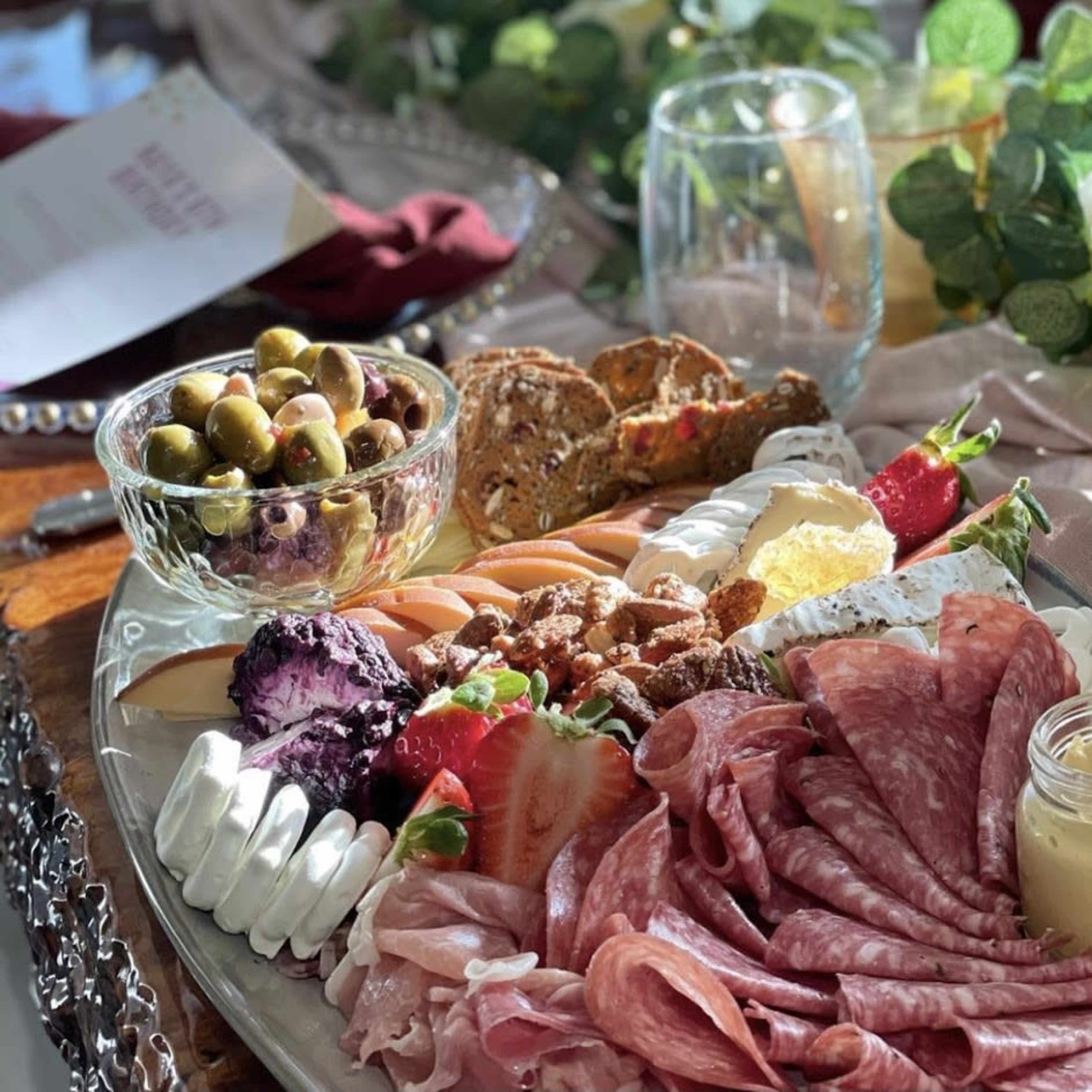 A large platter of assorted meats, cheeses, fruits, crackers, and olives arranged artfully on a wooden table surrounded by greenery and tableware.