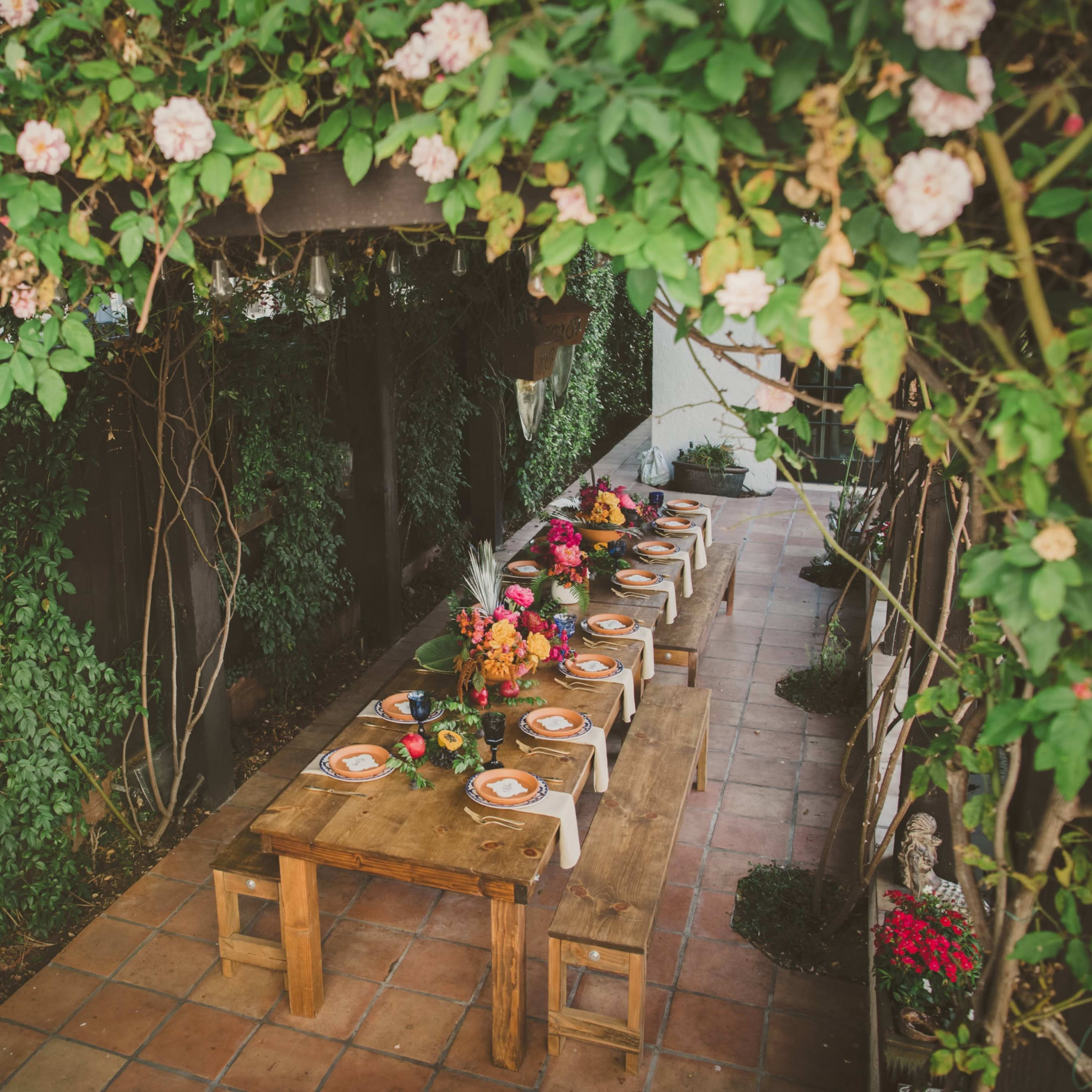 A long wooden table is set for a meal under a canopy of green foliage and blooming flowers.