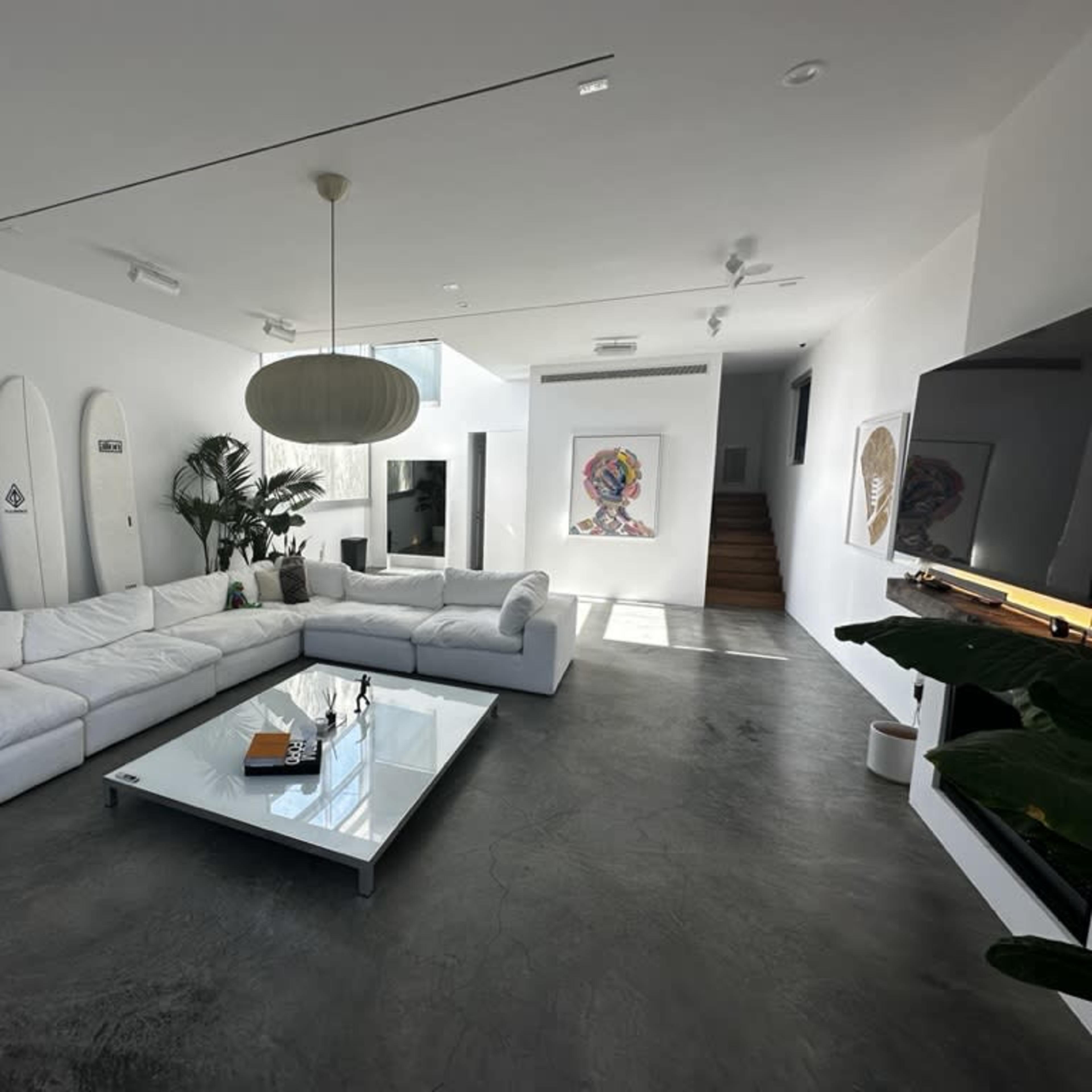 A modern living room with a large white sectional sofa, a glass coffee table, and indoor plants, set against a minimalist white wall and a staircase in the background.