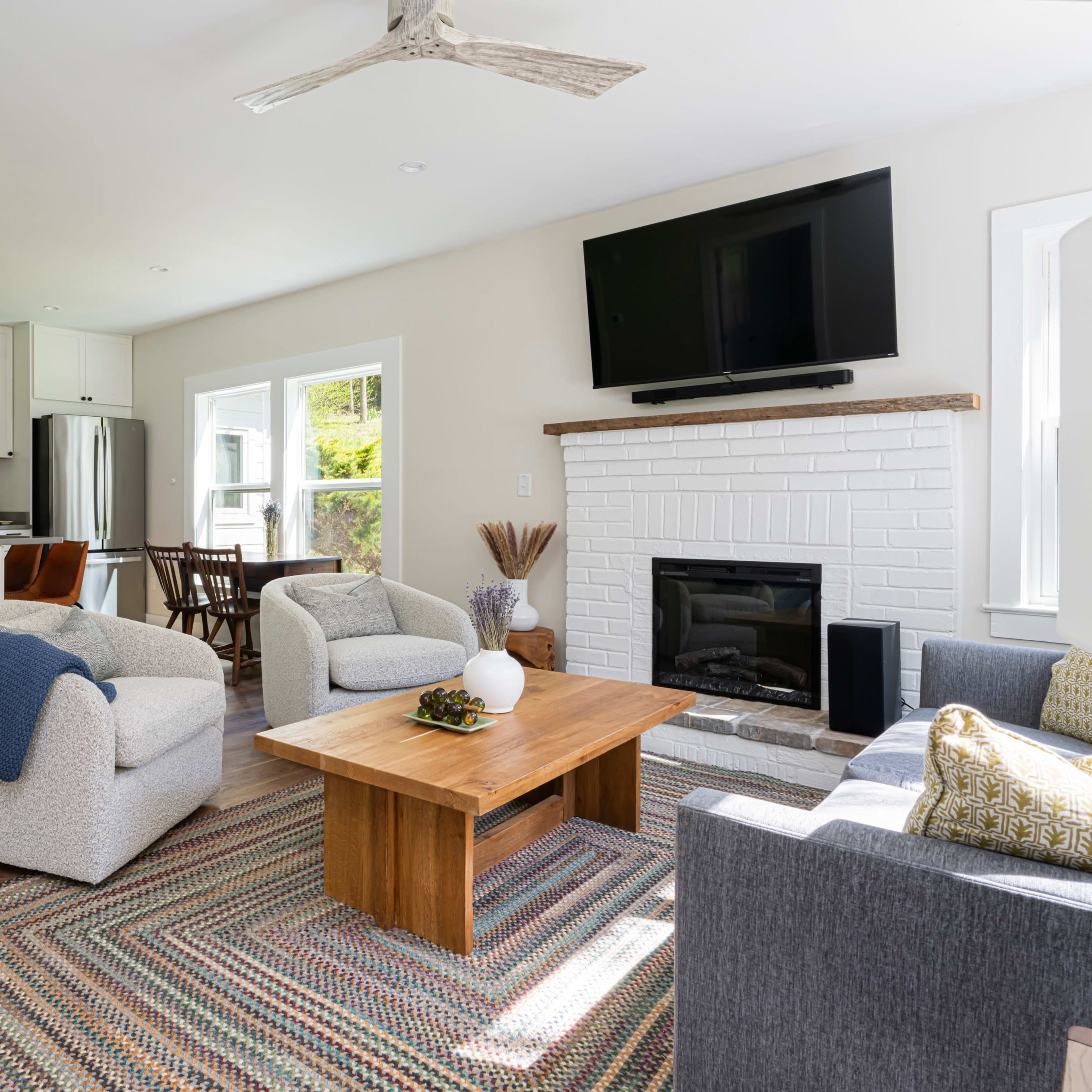 A modern living room features a fireplace, a coffee table, and comfortable seating, with an adjacent kitchen visible in the background.