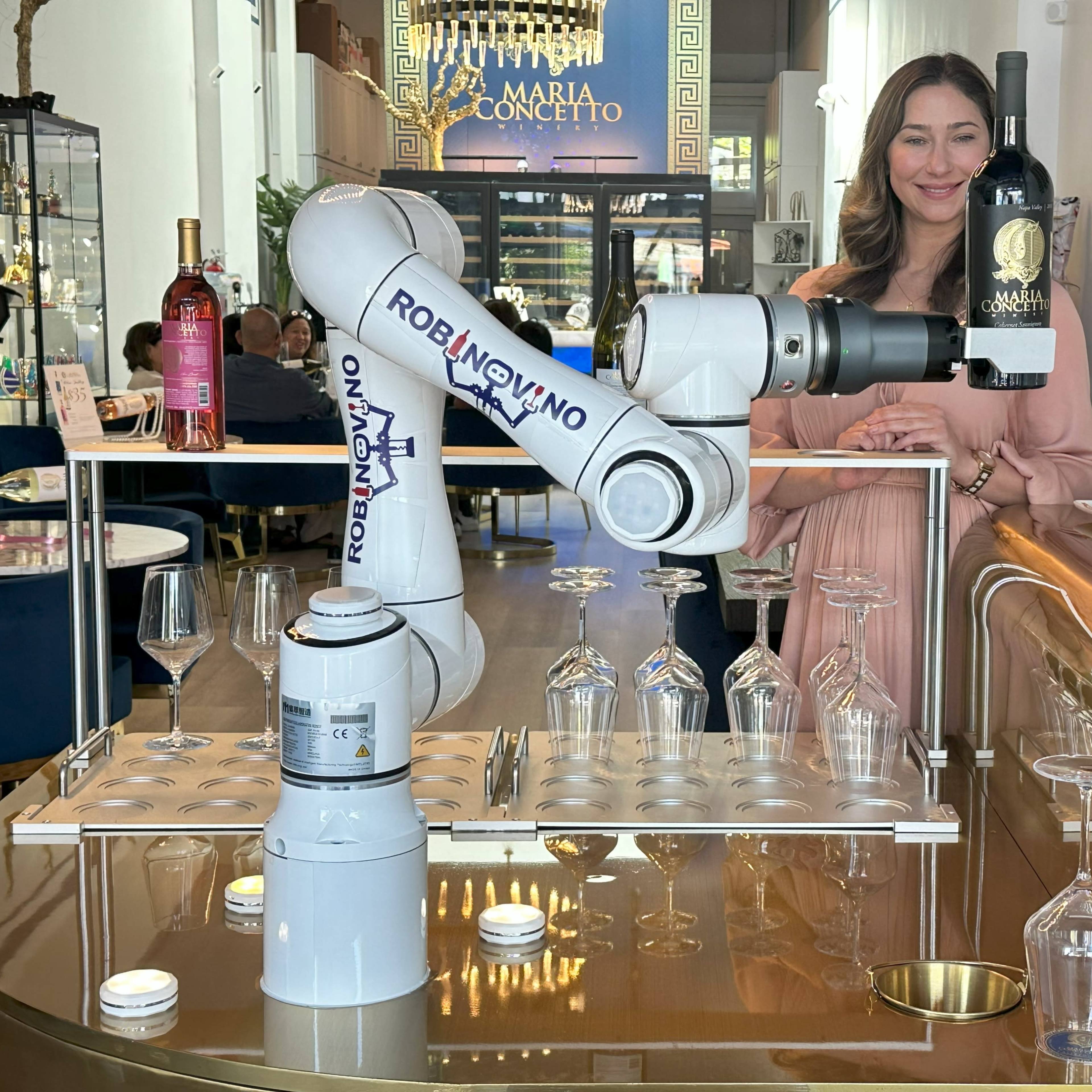 A woman stands behind a bar while a robotic arm serves wine bottles and prepares glasses.