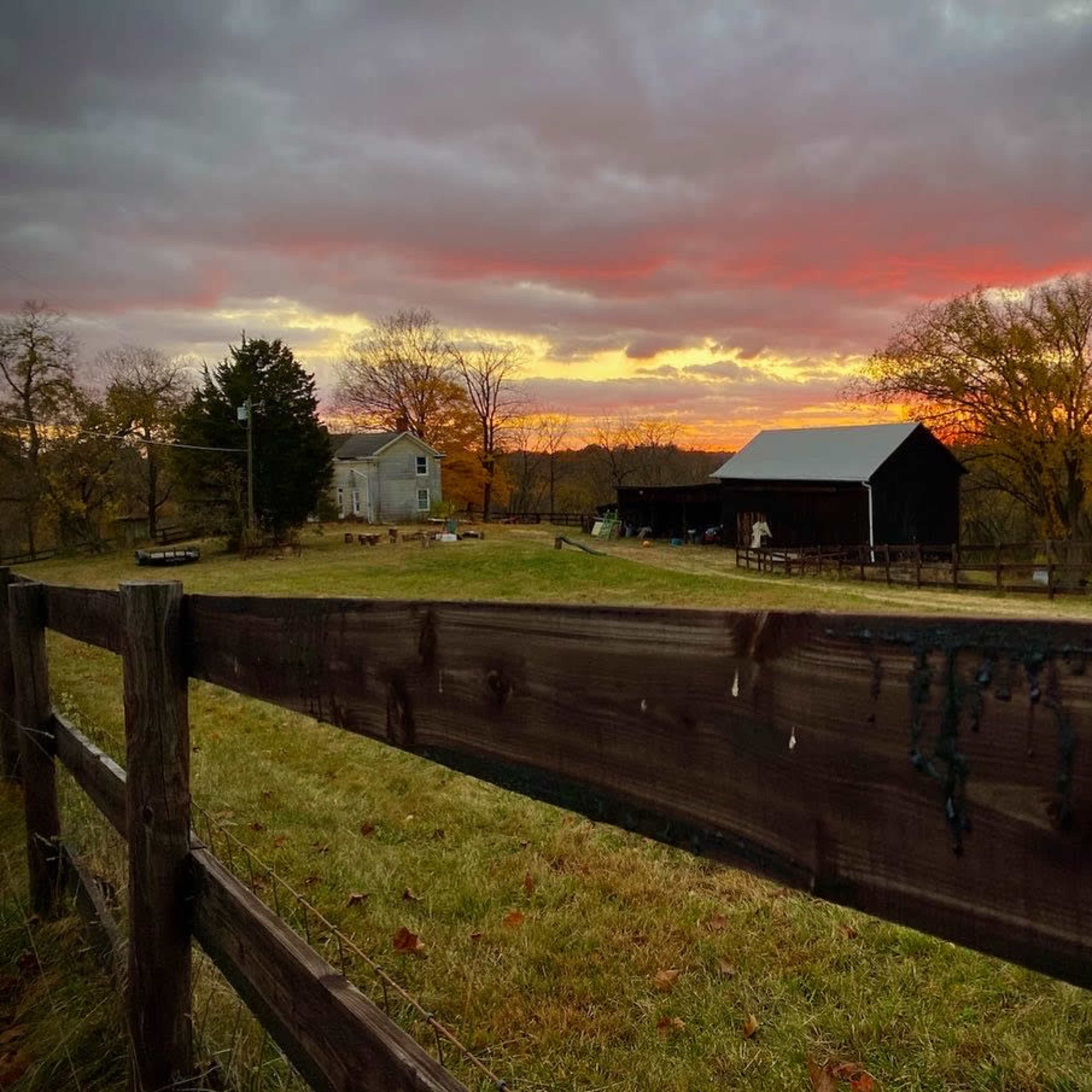A wooden fence lines a grassy field with a barn and a house silhouetted against a colorful sunset.