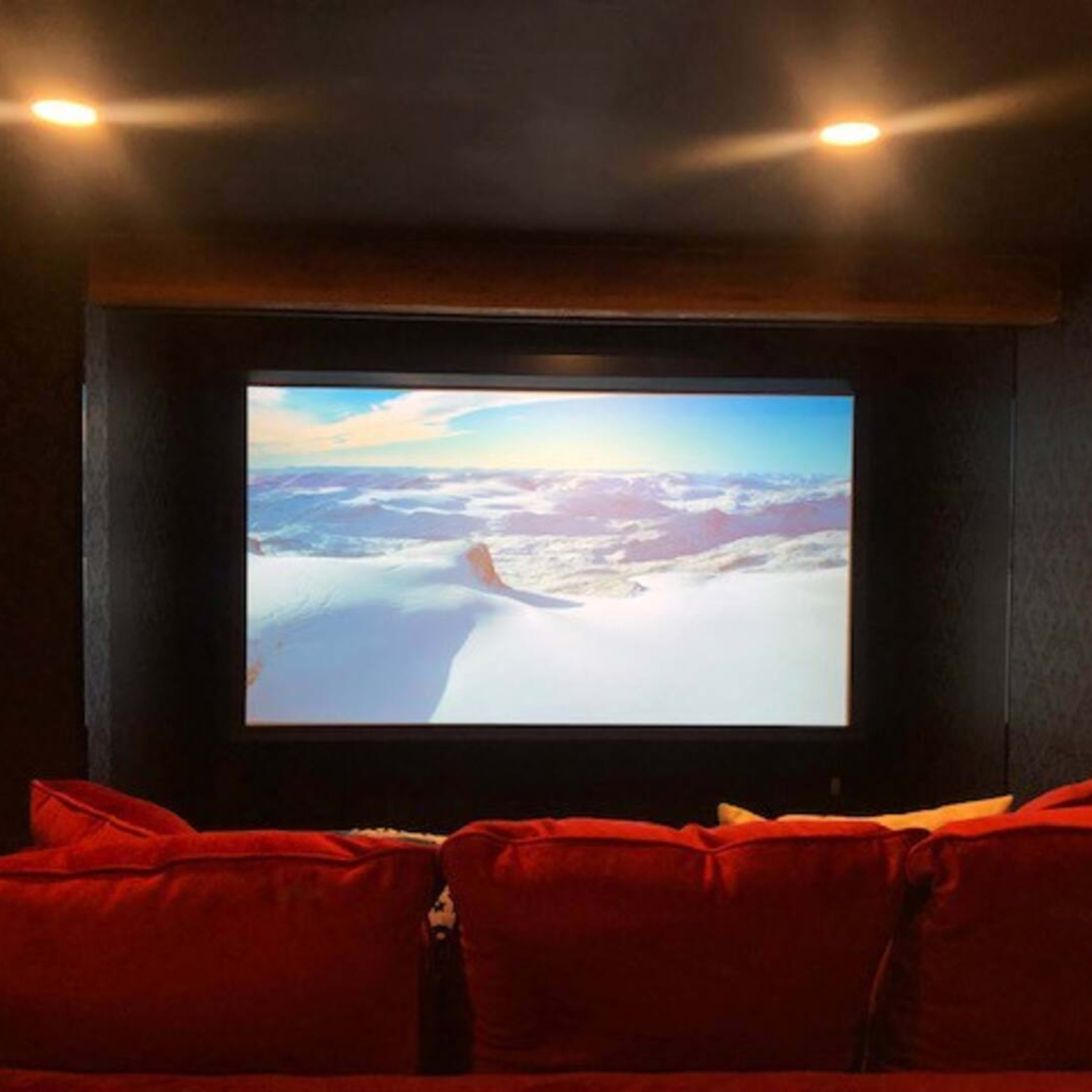 A large projection screen displaying a snowy mountain landscape is set against a dark wall in a cozy home theater with red seating.
