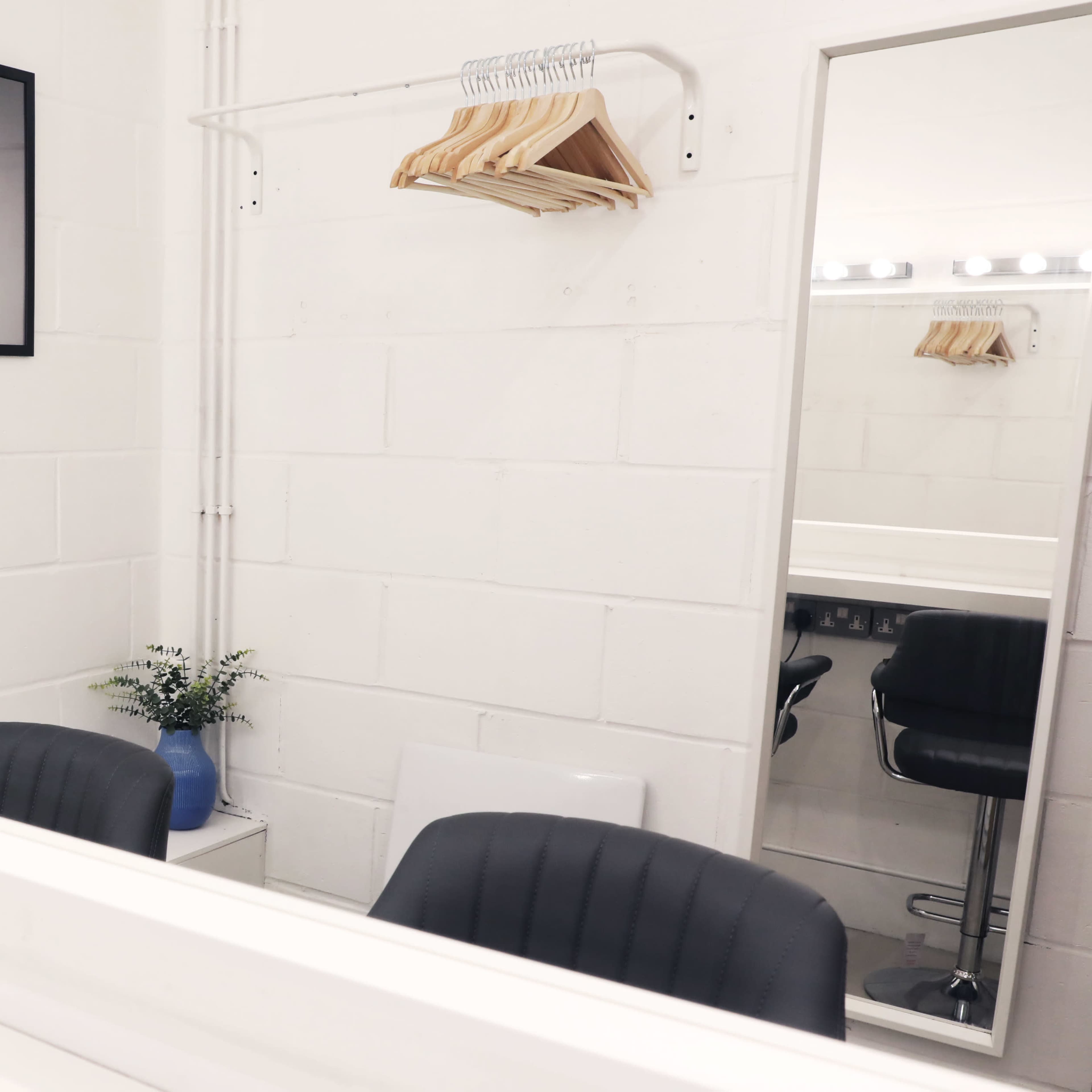 A minimalistic dressing room with two black chairs, a mirror, and framed artworks on the wall, along with a small vase of greenery on a table.