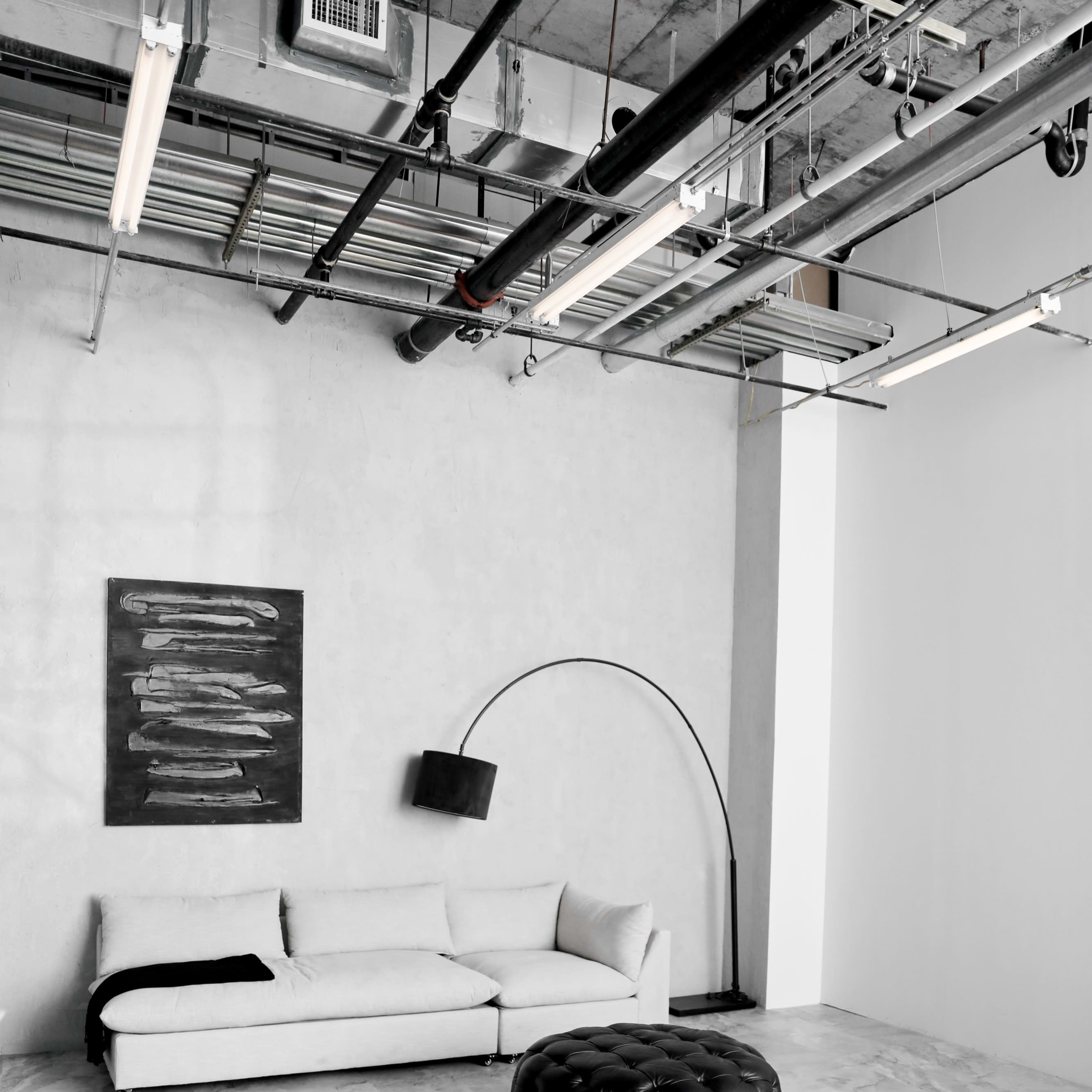 A minimalist living space with a gray wall, a white sofa, a black floor lamp, and a black tufted ottoman on a marble floor.