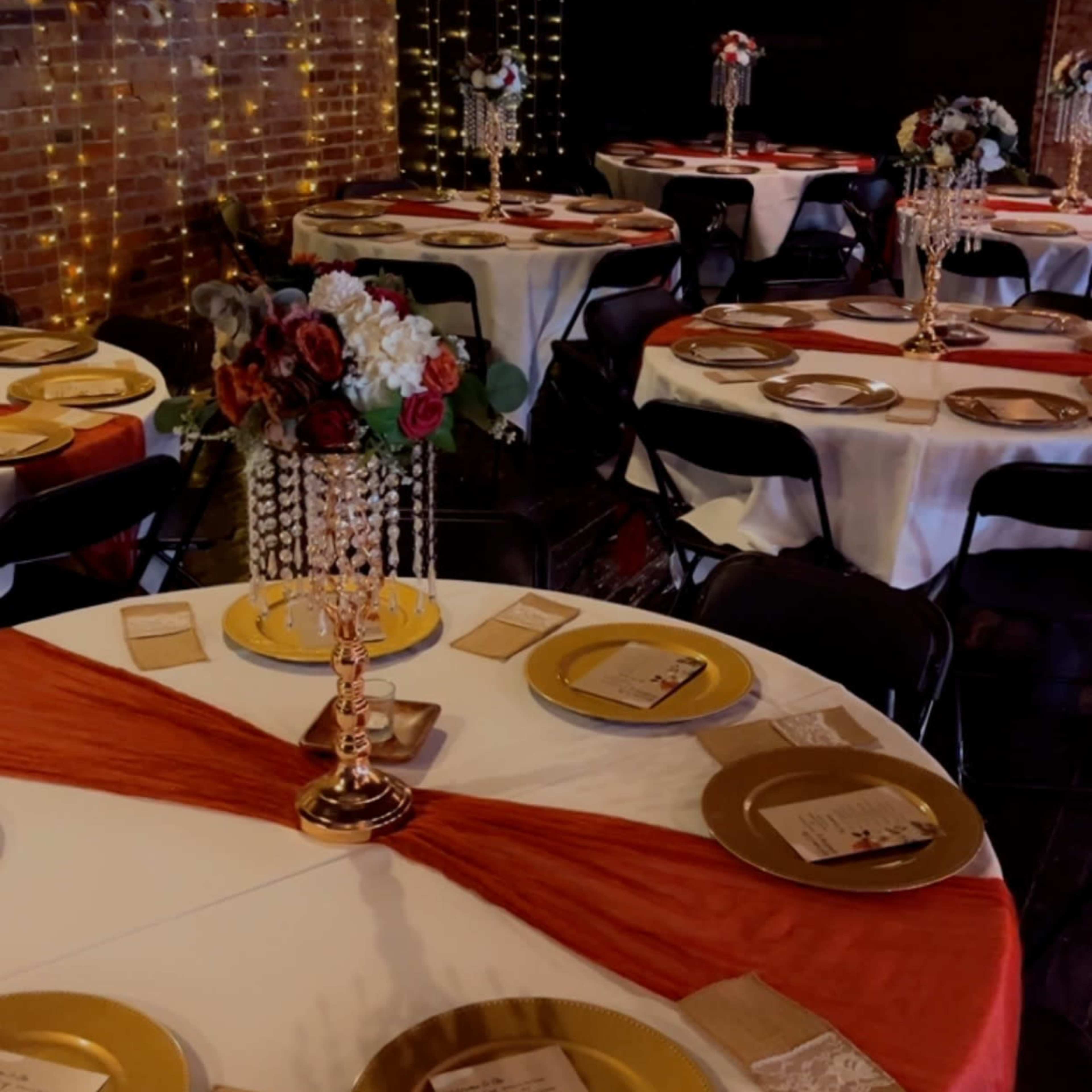 A dining area is set with white and orange table linens, gold plates, and floral centerpieces under soft lighting against a brick wall.