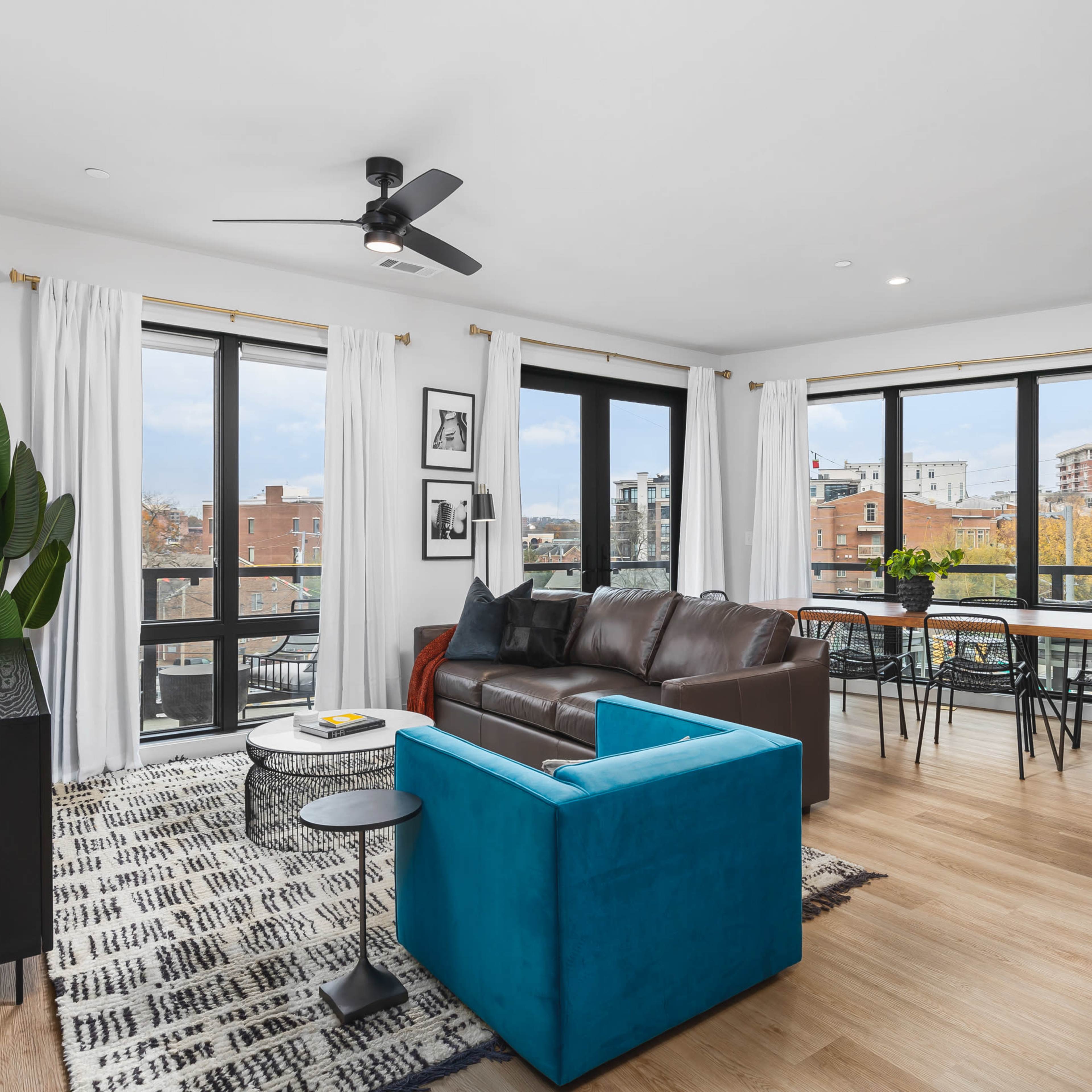 A contemporary living space features a brown leather sofa, a blue accent chair, and large windows overlooking an urban landscape.