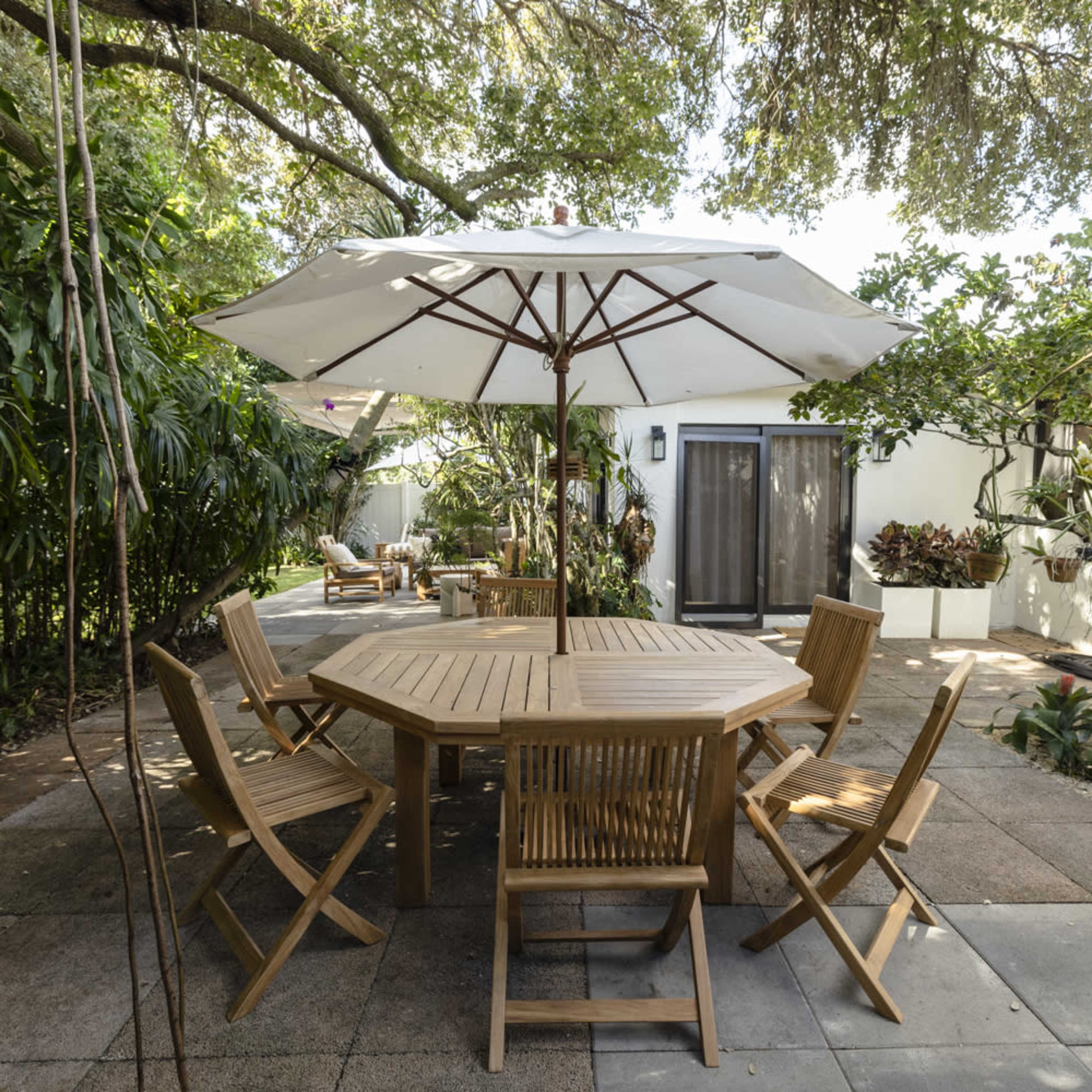 A wooden outdoor dining table is surrounded by six folding chairs under a large umbrella in a garden setting with greenery and a white building in the background.