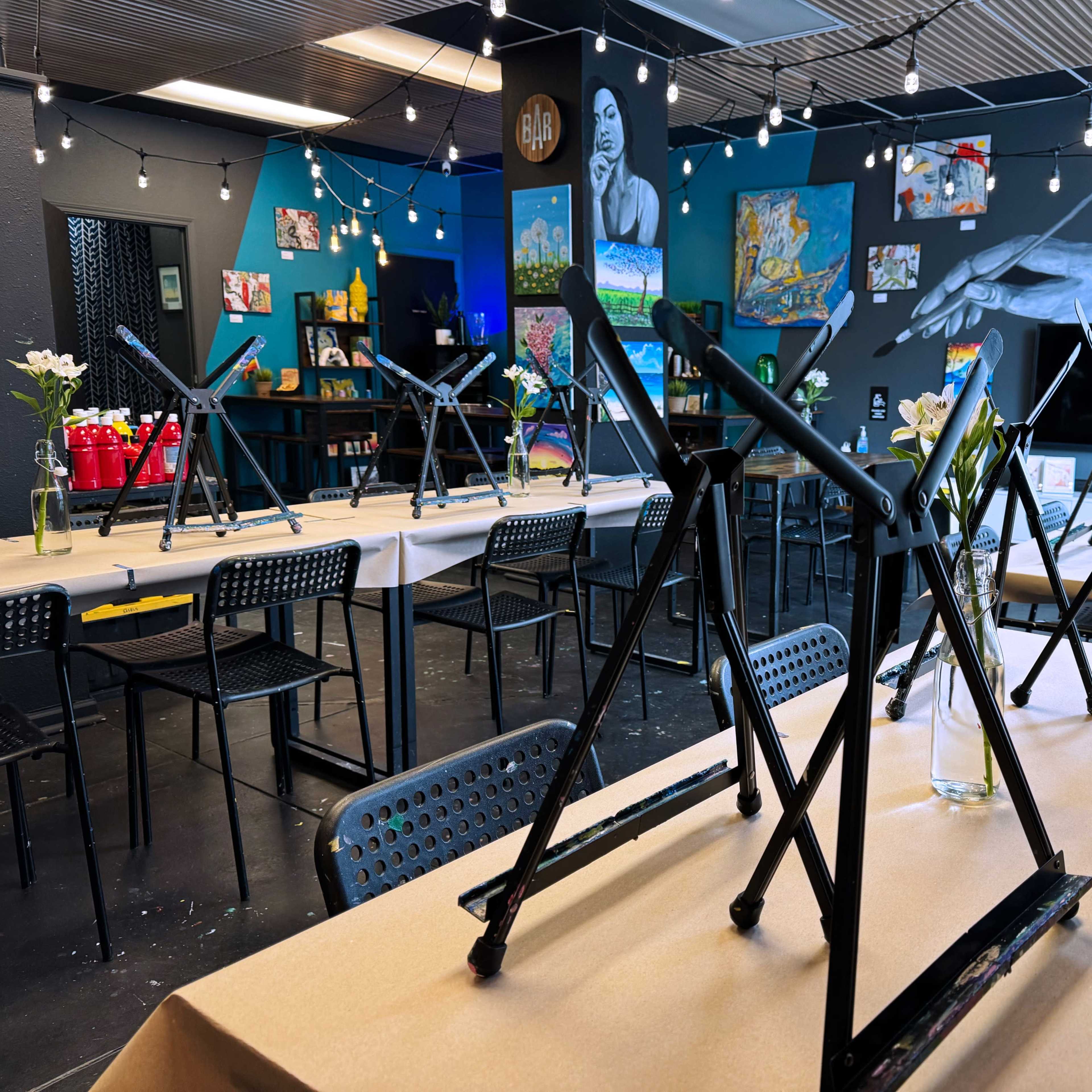 An art studio is set up with tables covered in brown paper, each equipped with an easel and surrounded by string lights and artworks on the walls.