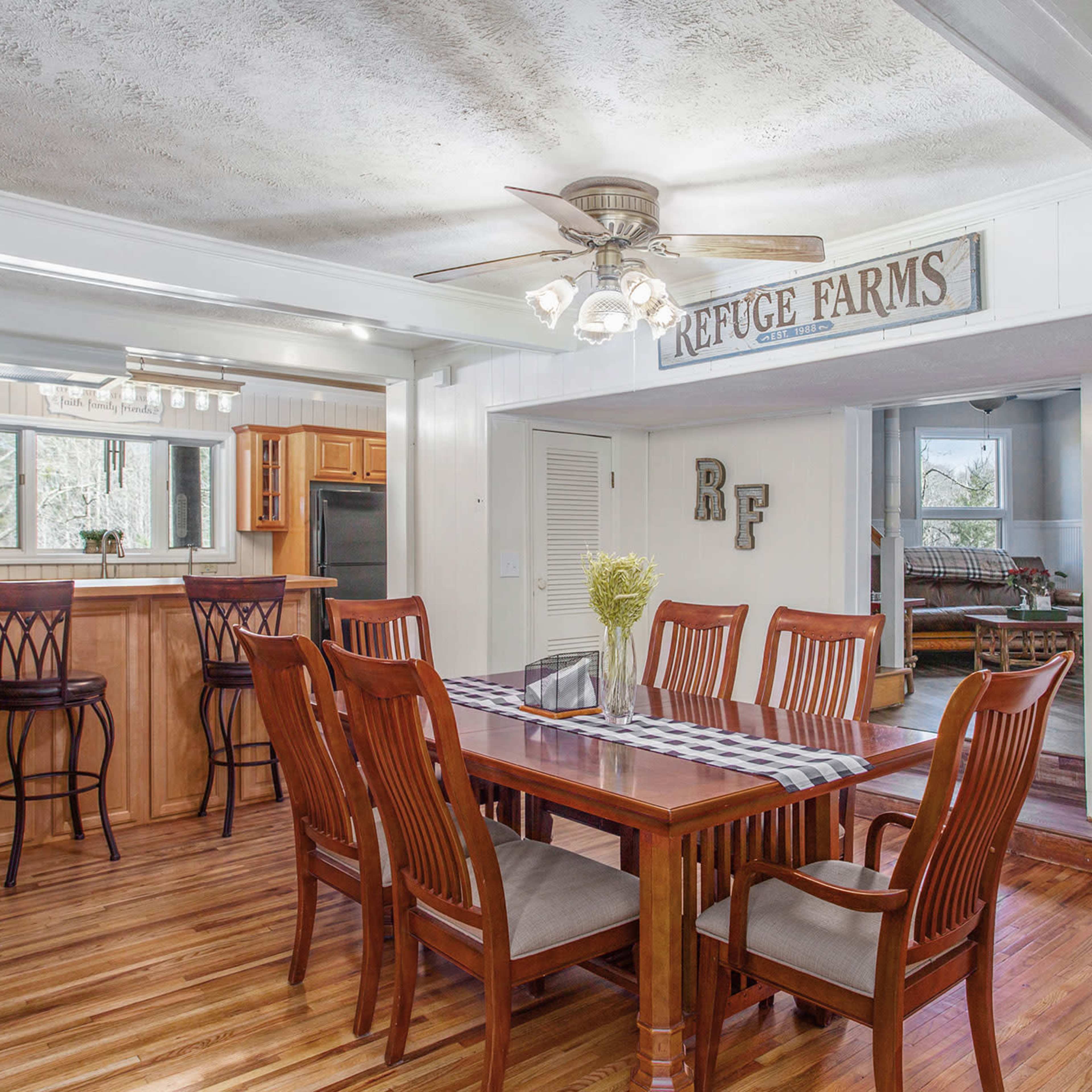 A dining area with a wooden table surrounded by chairs, adjacent to a kitchen and living space, illuminated by a ceiling fan and natural light.