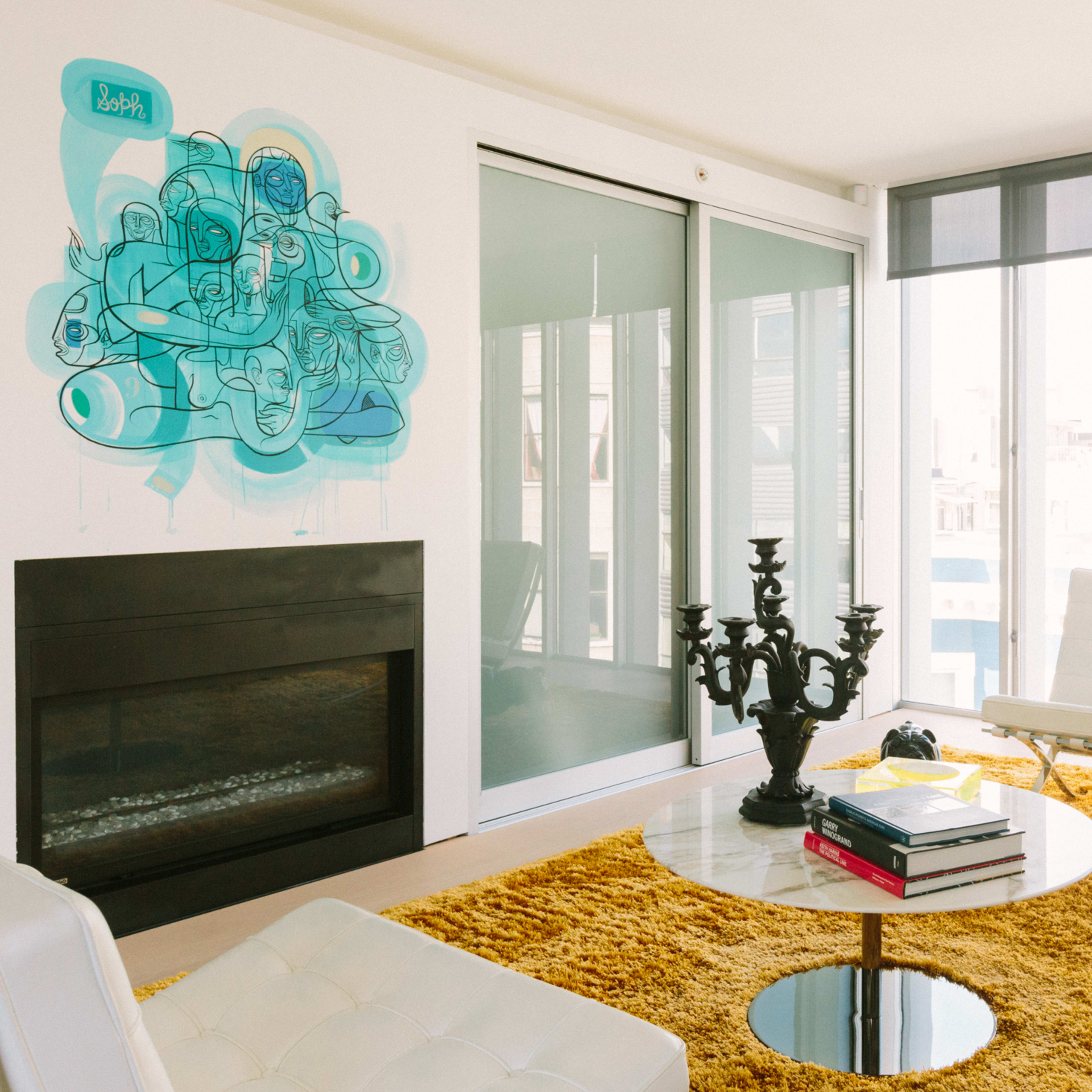 A modern living room features a white sofa, a circular glass coffee table on a yellow area rug, a black candelabra, and large windows allowing natural light to enter.