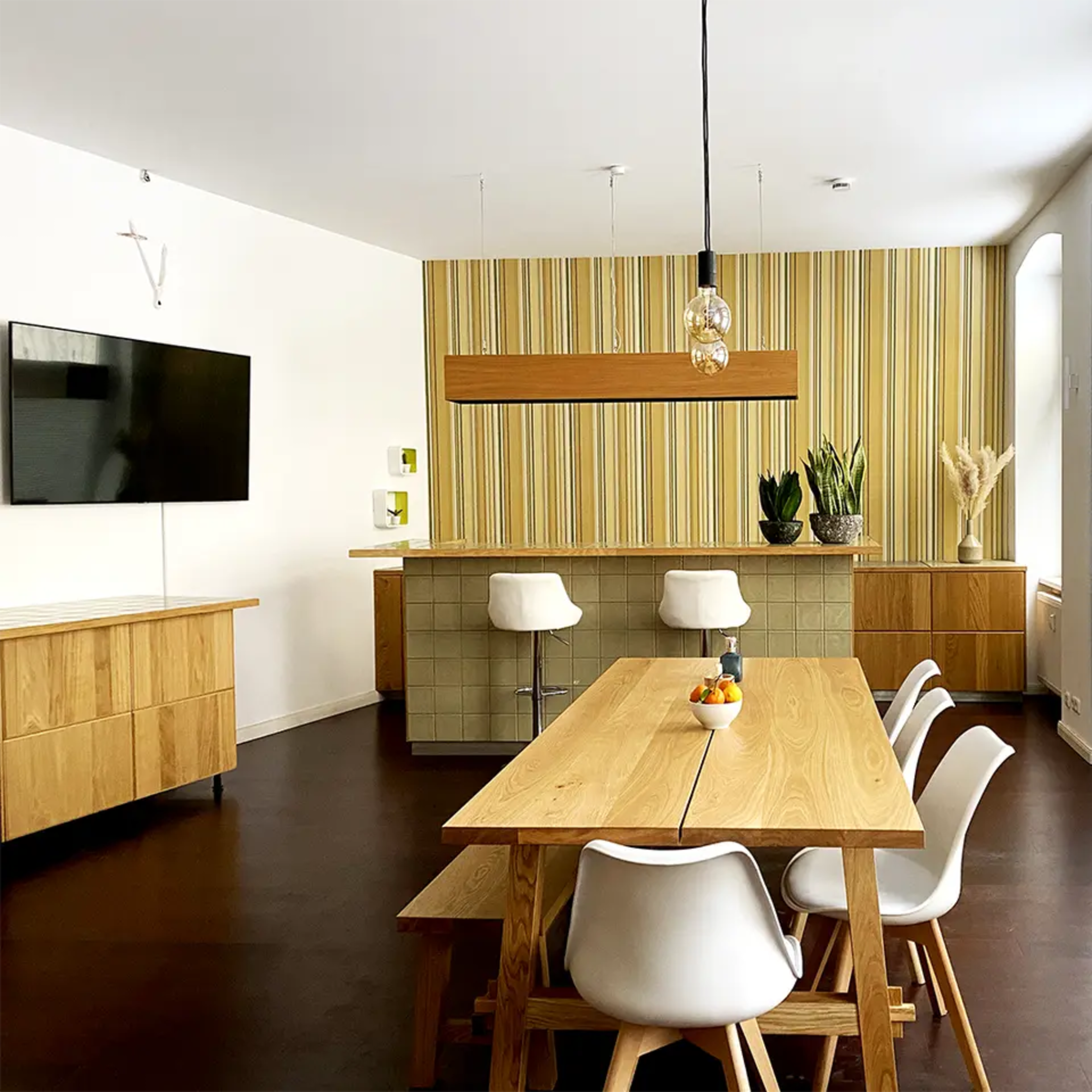 A modern dining area featuring a long wooden table, white chairs, and a kitchen bar with a striped wall design.