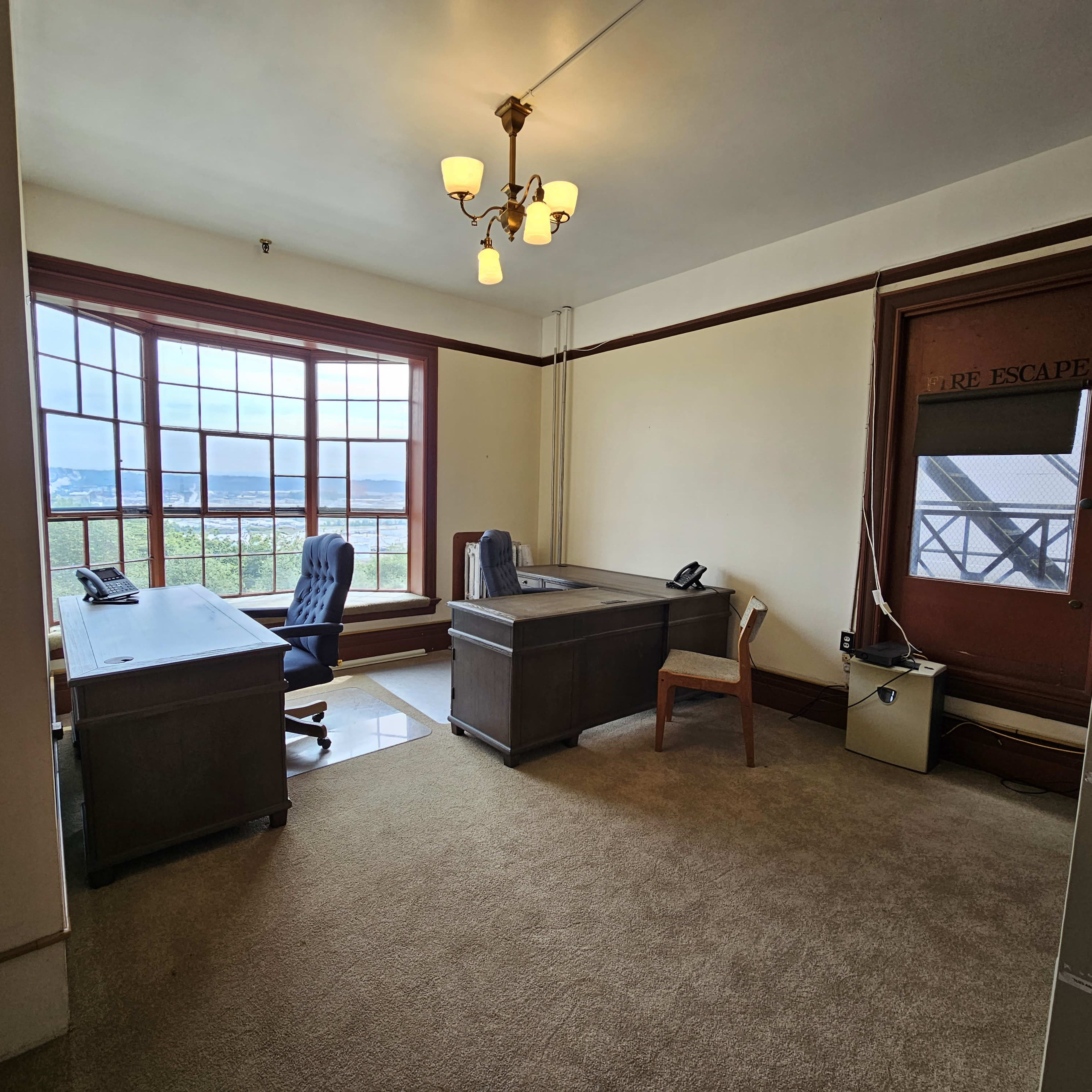 A shared office space features two desks, a chair, and a large window with views of the outdoors.