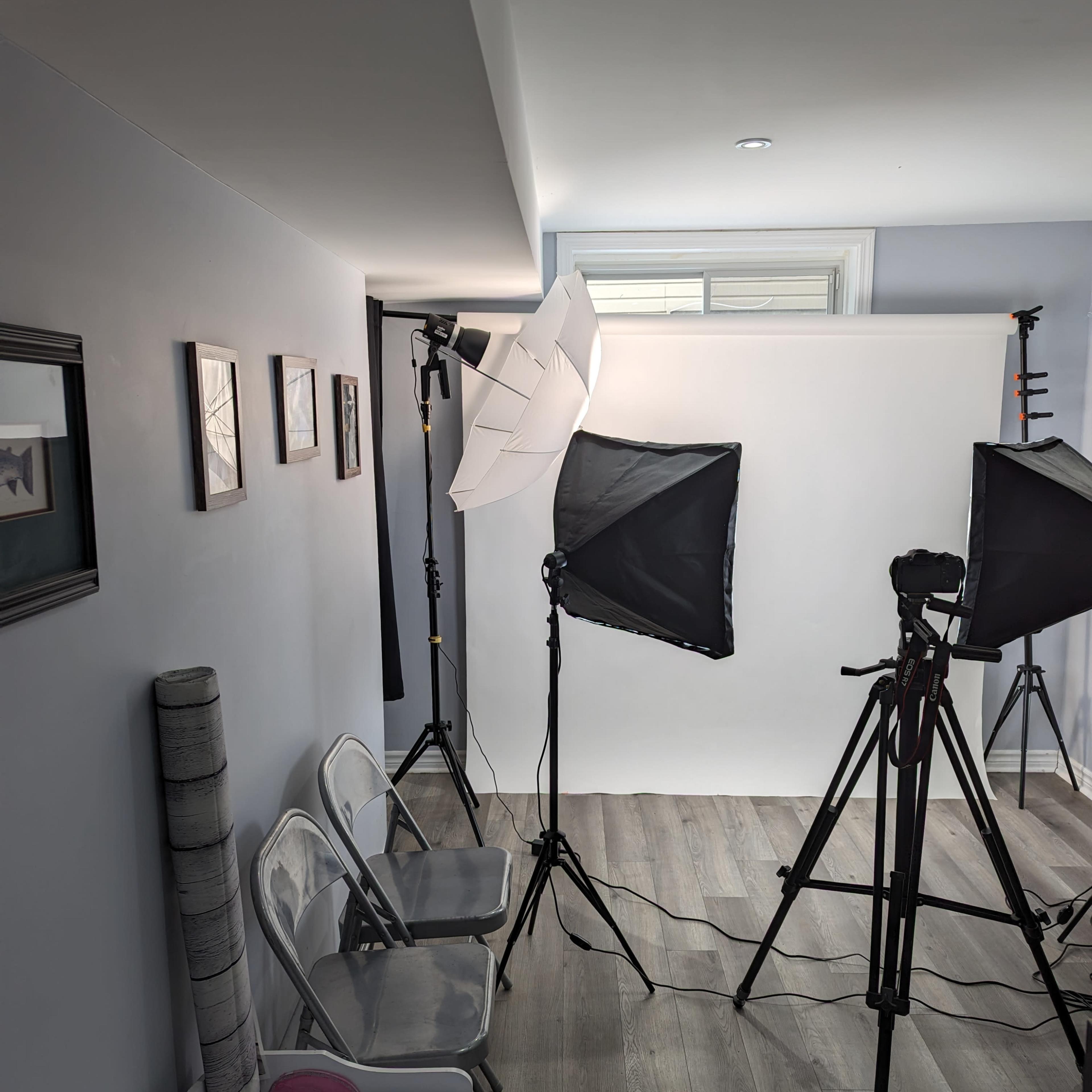 A photography studio setup with two light umbrellas, tripods, and empty chairs against a gray wall.