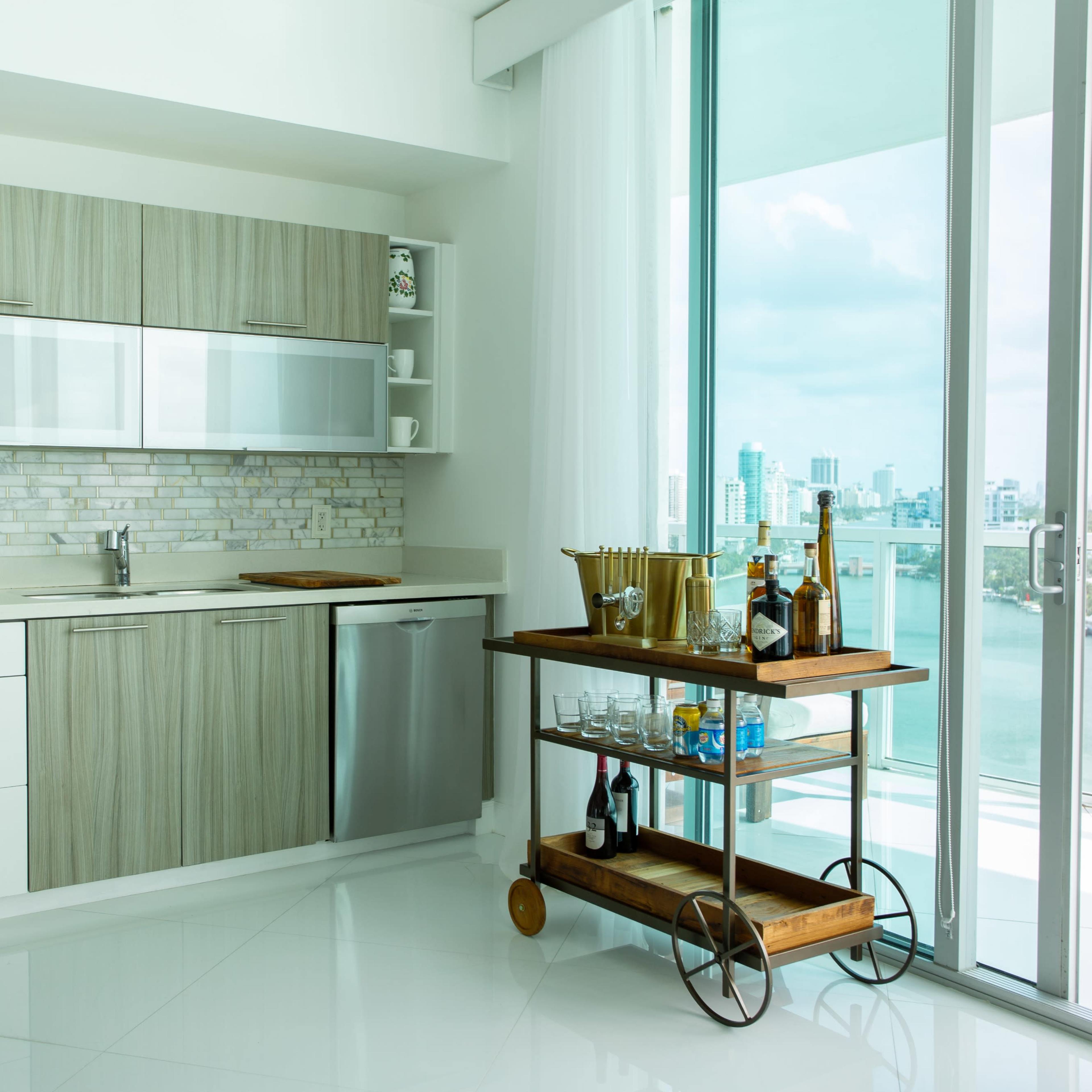 A modern kitchen features sleek cabinetry, stainless steel appliances, and a wooden bar cart with drinks, set against a backdrop of large windows overlooking a waterfront city view.