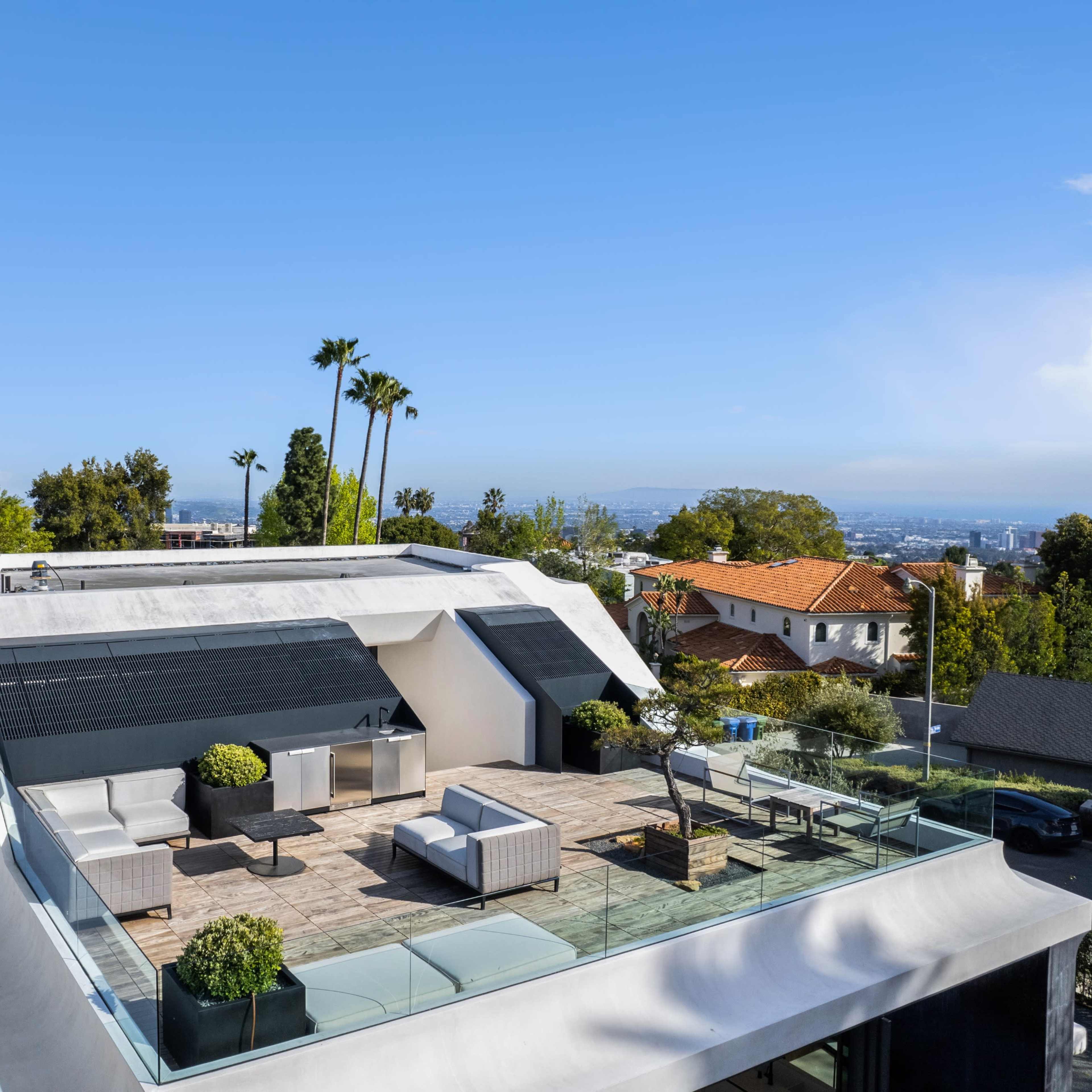 A modern rooftop terrace with outdoor furniture, surrounded by greenery and overlooking a cityscape.