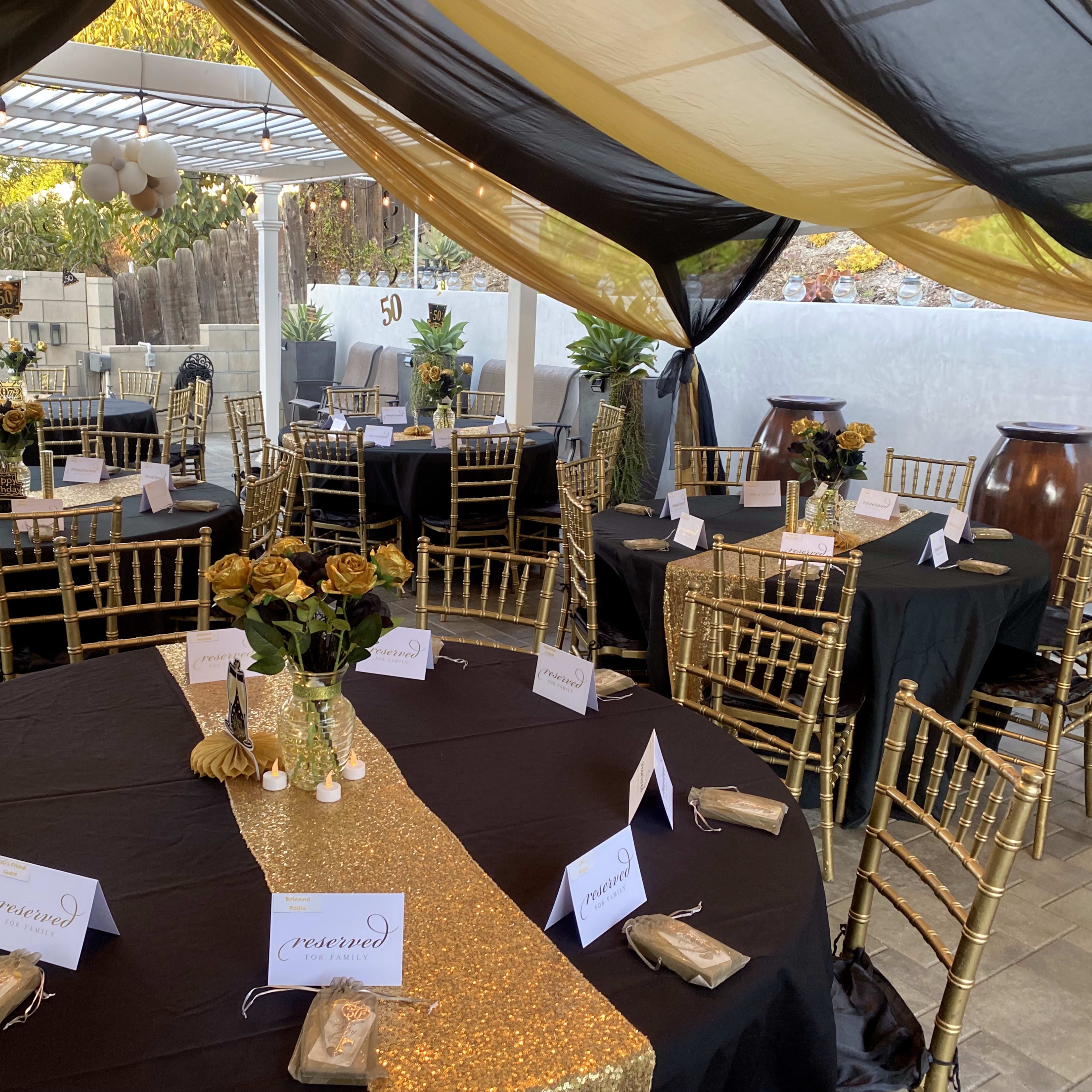 A decorated outdoor event space features round tables covered with black tablecloths, adorned with gold centerpieces and place cards, under a black and gold canopy.