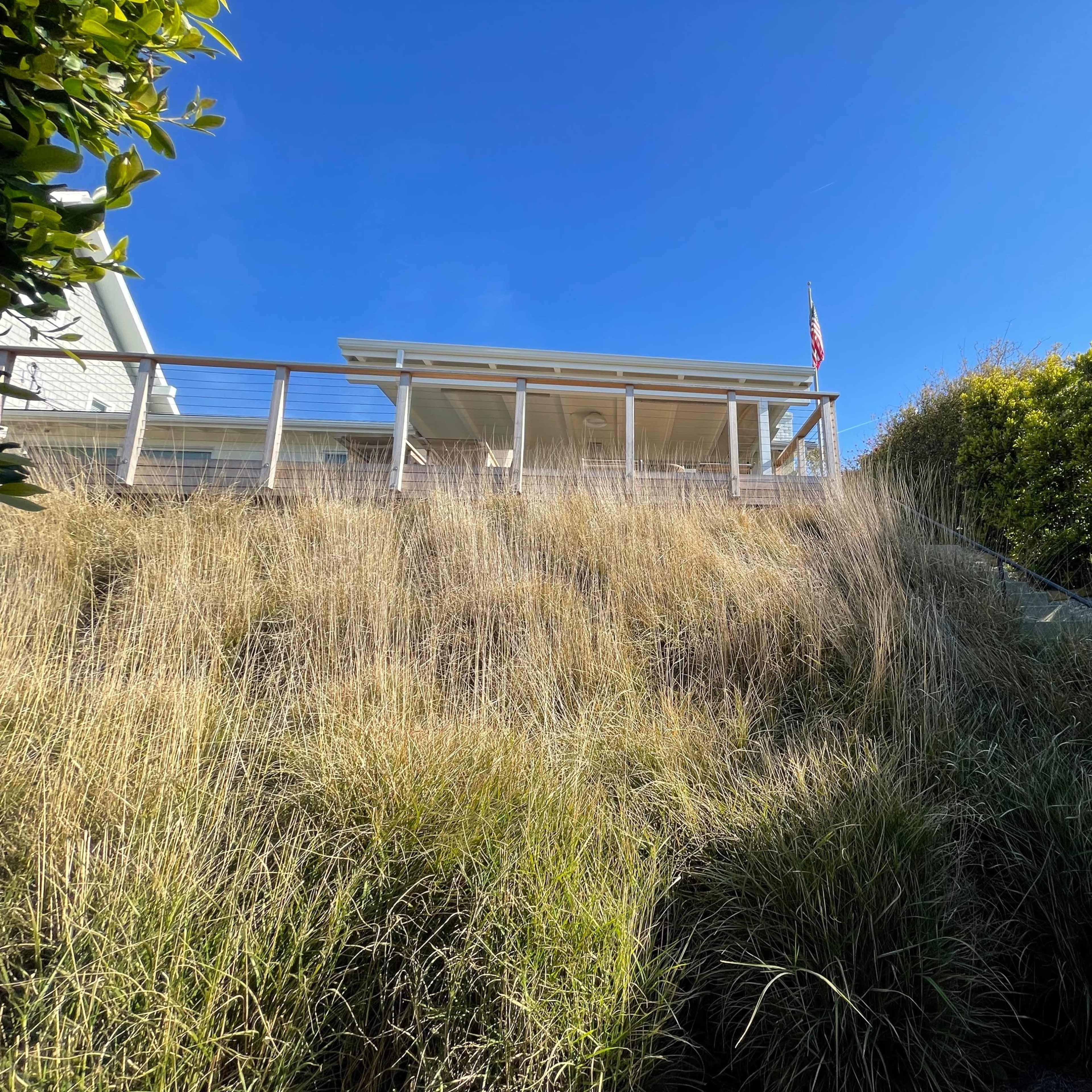 A house with a porch sits atop a grassy hill, with a flag visible and a set of stairs leading up from the foreground.