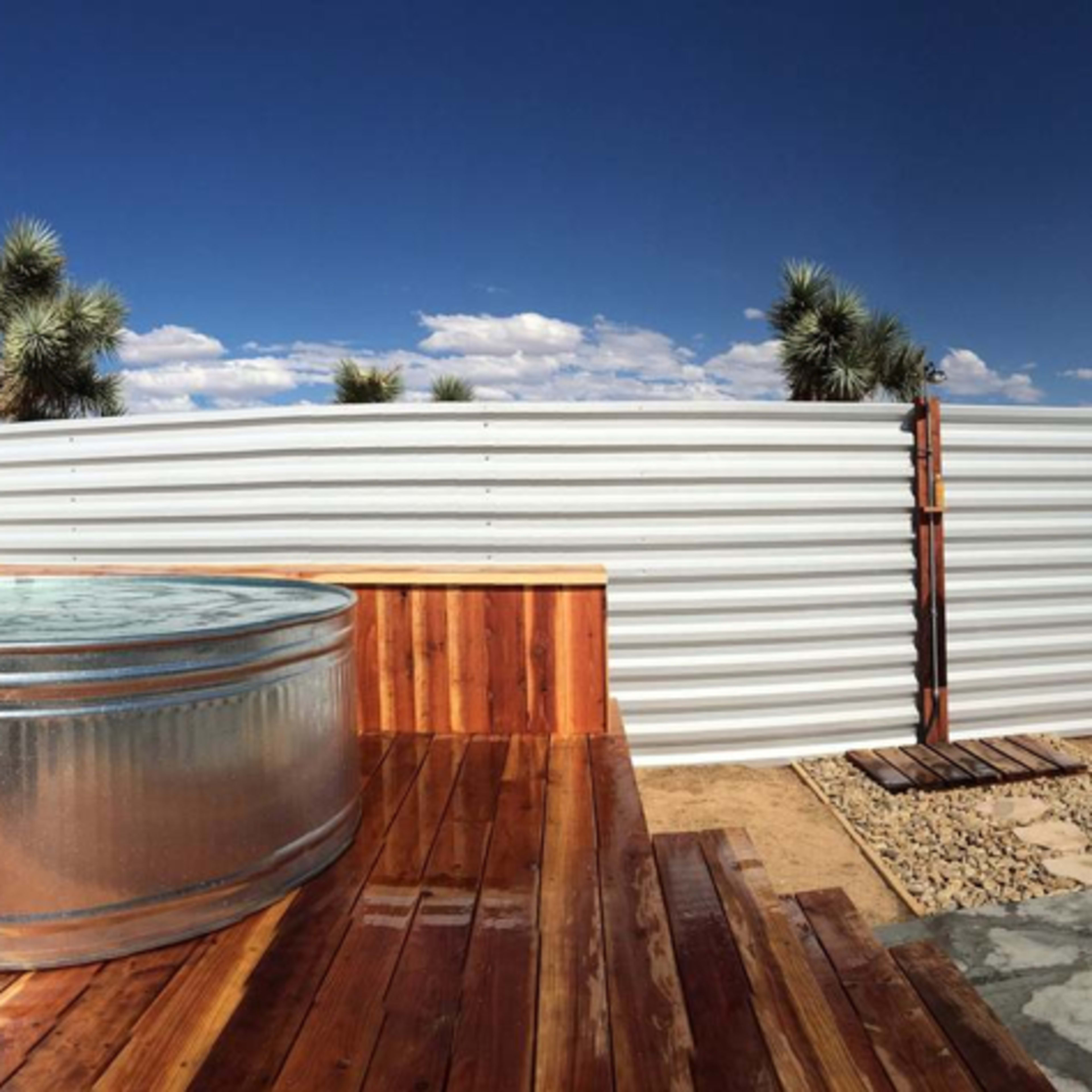A circular metal tub filled with water sits on a wooden deck surrounded by corrugated metal fencing and desert vegetation under a blue sky.