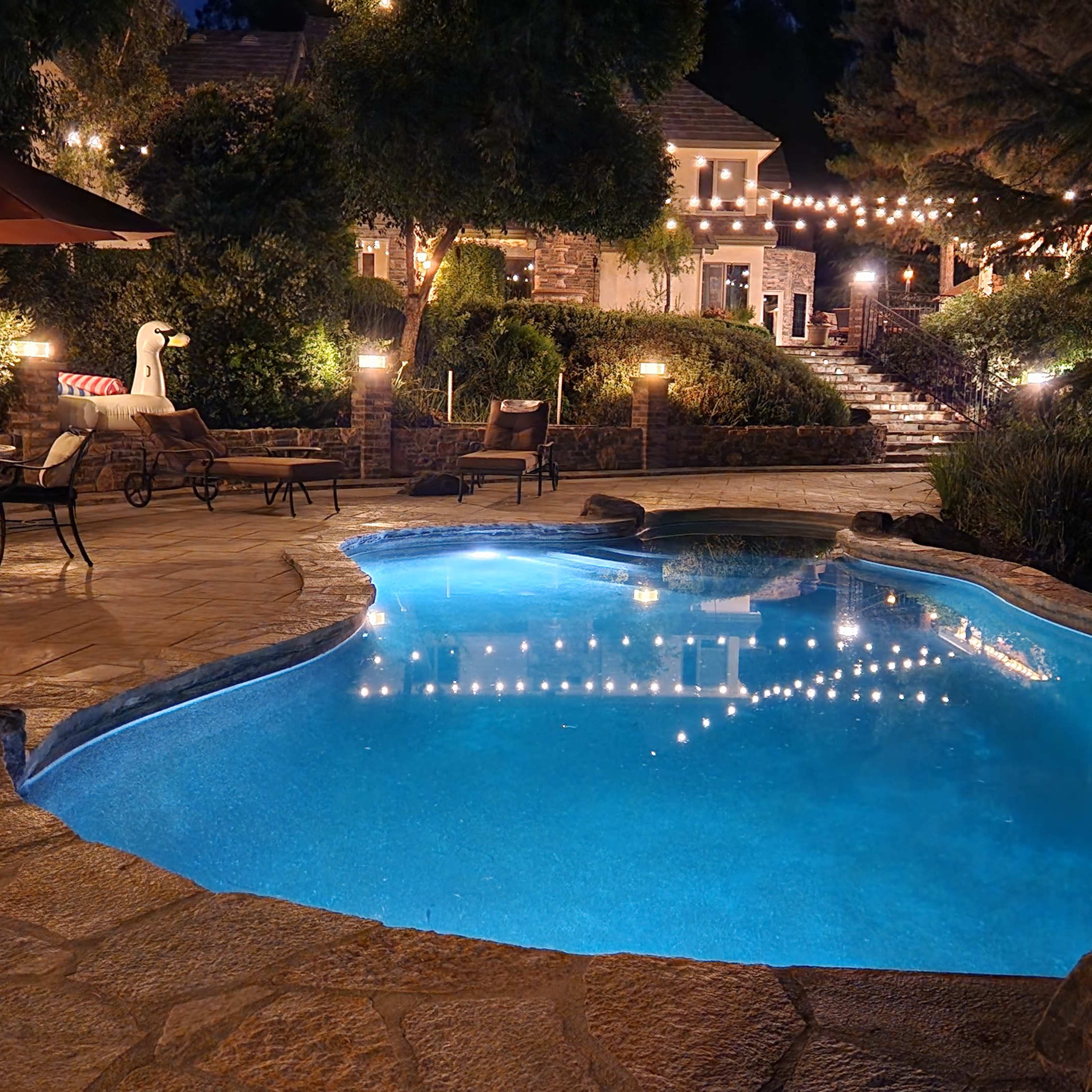 A beautifully lit pool area features a curved pool surrounded by stone paving, lounge chairs, and decorative lighting in a residential backyard.