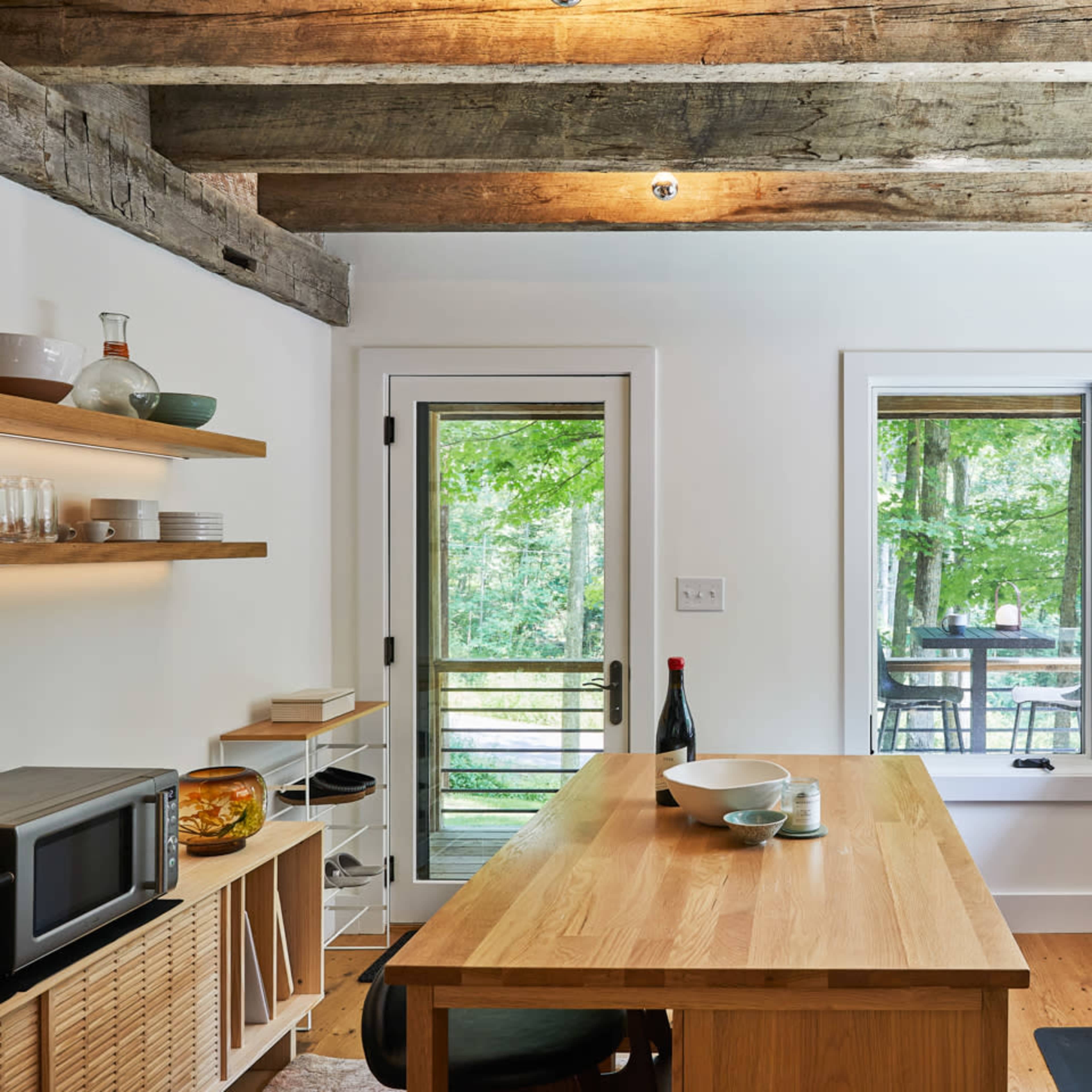 A modern kitchen features wooden beams, an island table, open shelving, and views of a forested outdoor deck through large windows.