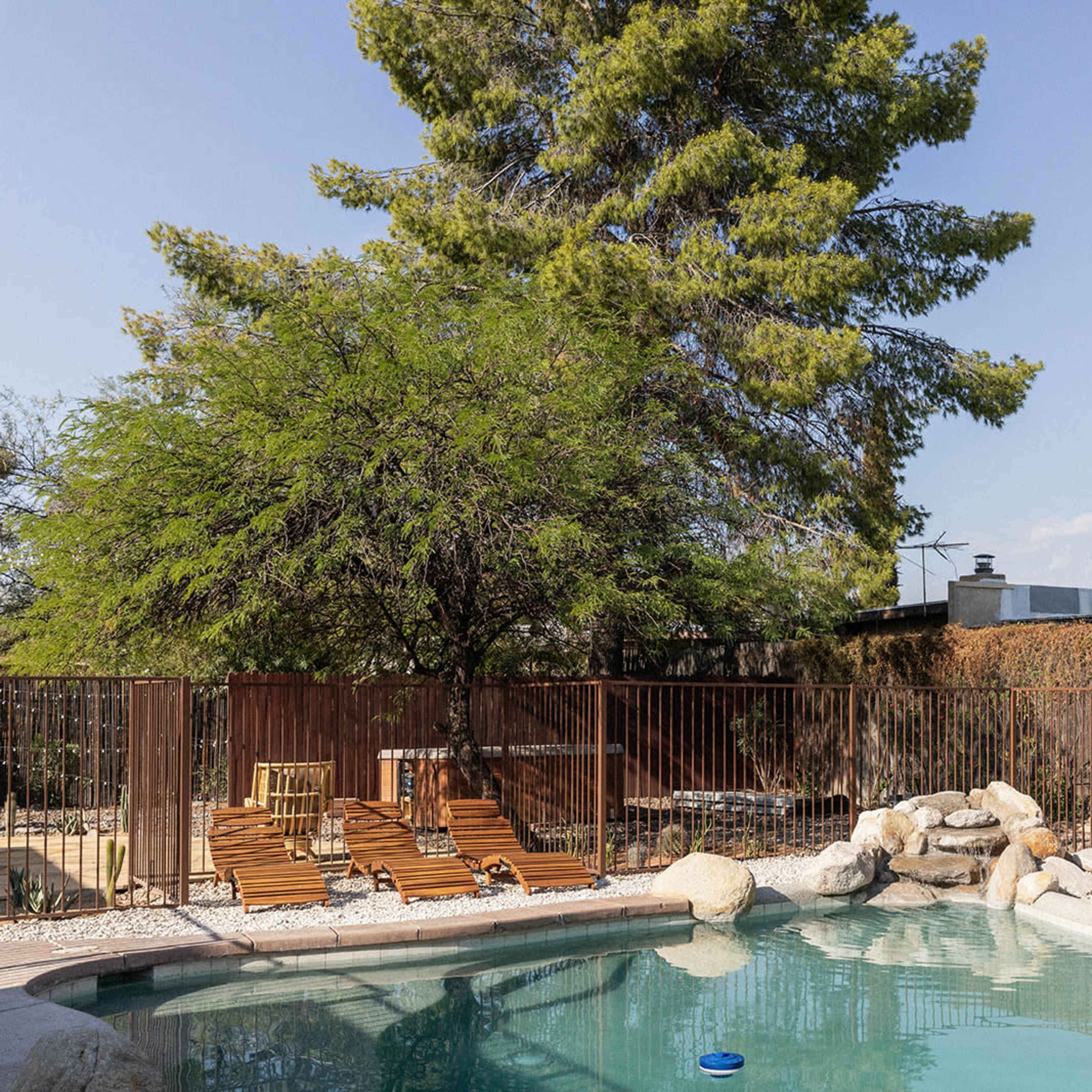 A swimming pool with a rock feature is surrounded by a metal fence, sun loungers, and trees in a residential backyard.