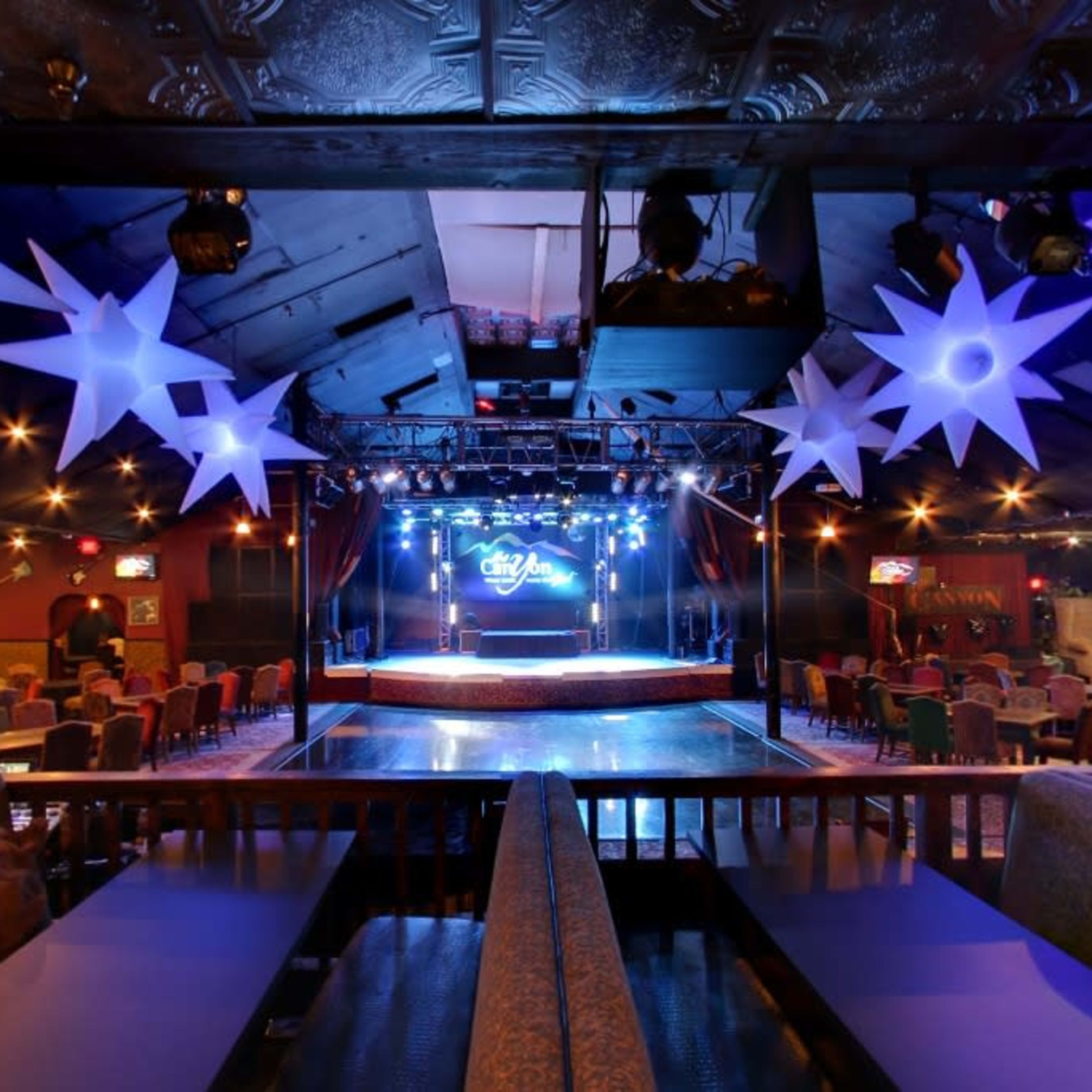 A spacious entertainment venue with a stage, illuminated by colorful lights, surrounded by seating areas and decorations.