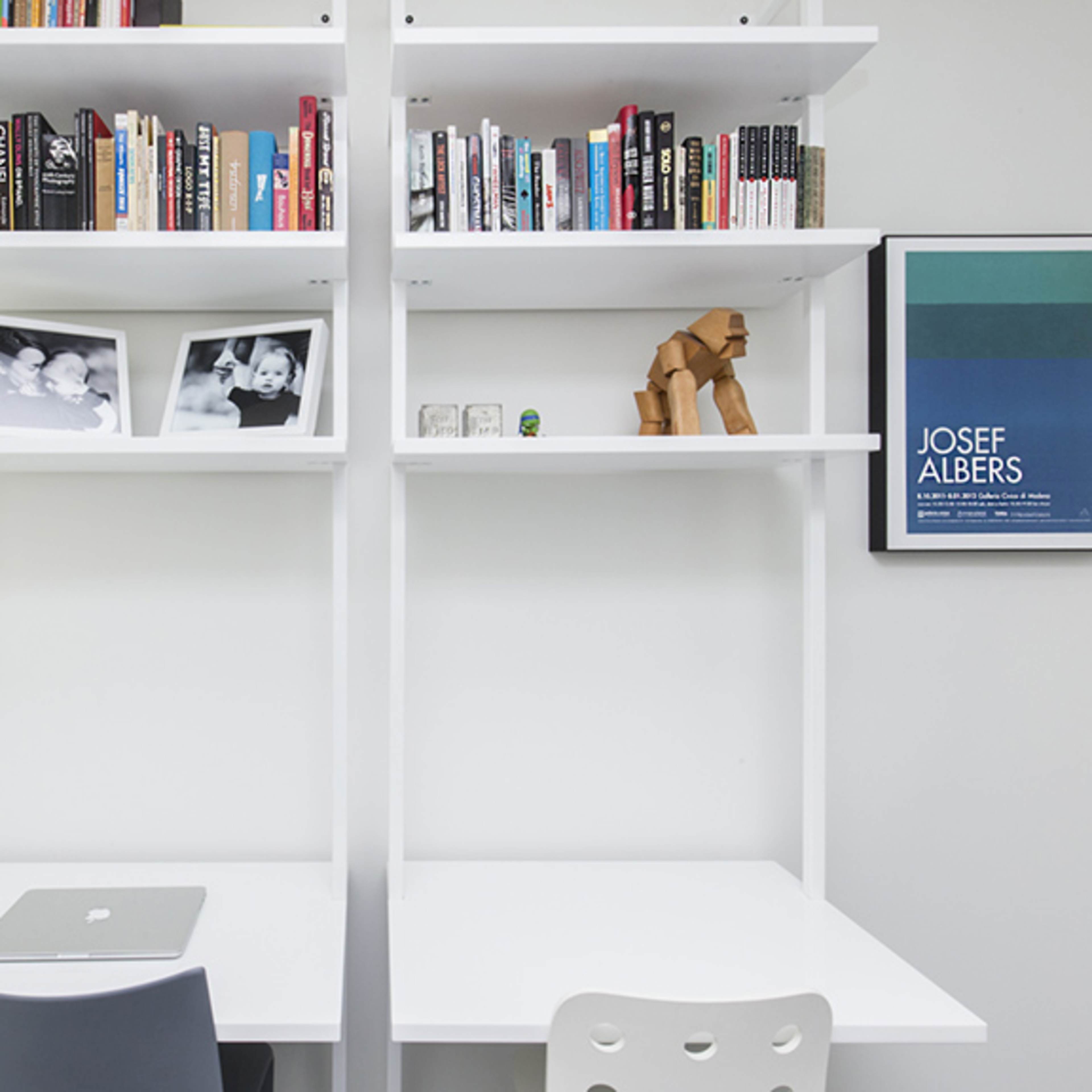 A minimalist workspace with two white desks, a bookshelf filled with books, and framed photographs on the wall.