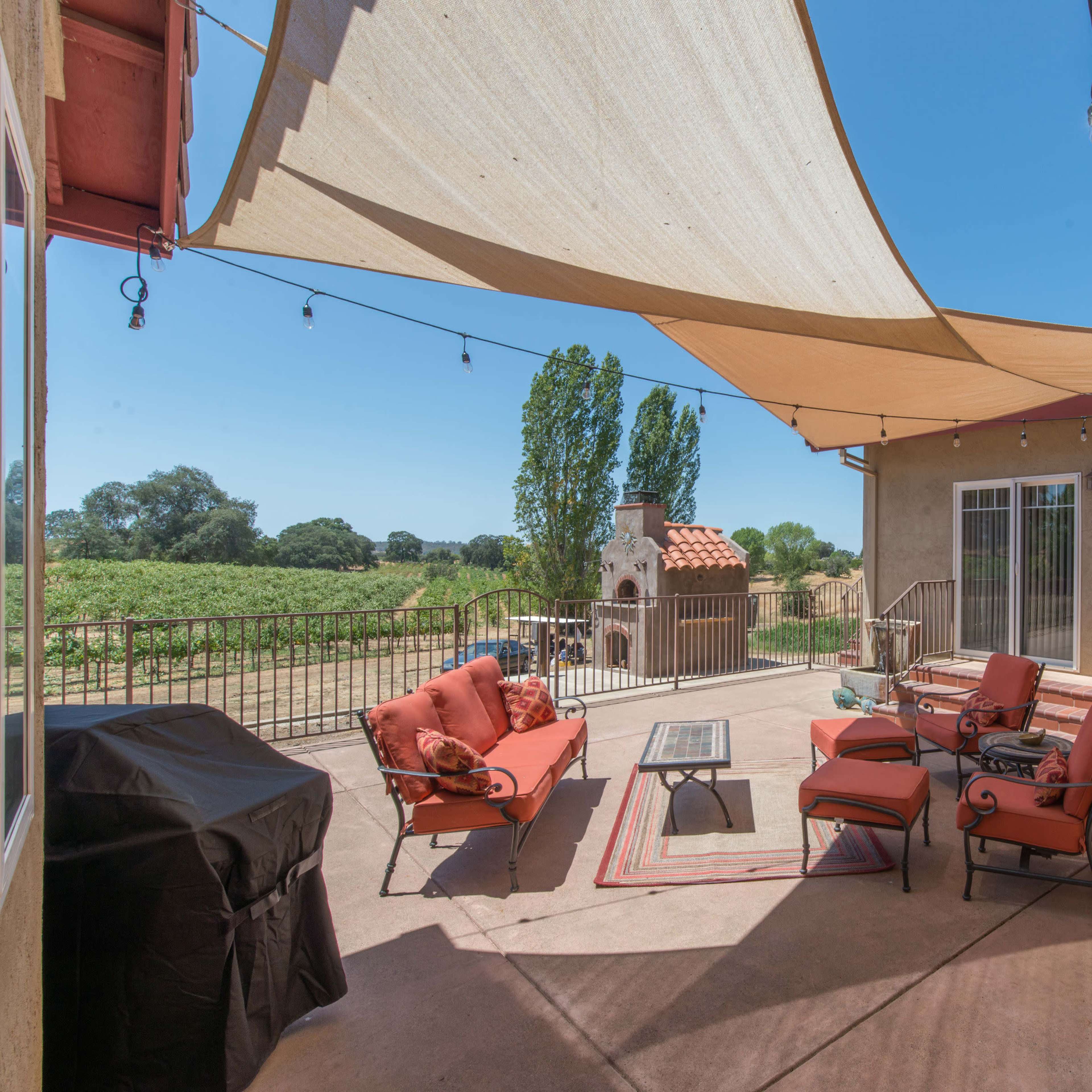 A patio area featuring red cushioned furniture, a barbecue grill, and a view of surrounding greenery under a shade canopy.
